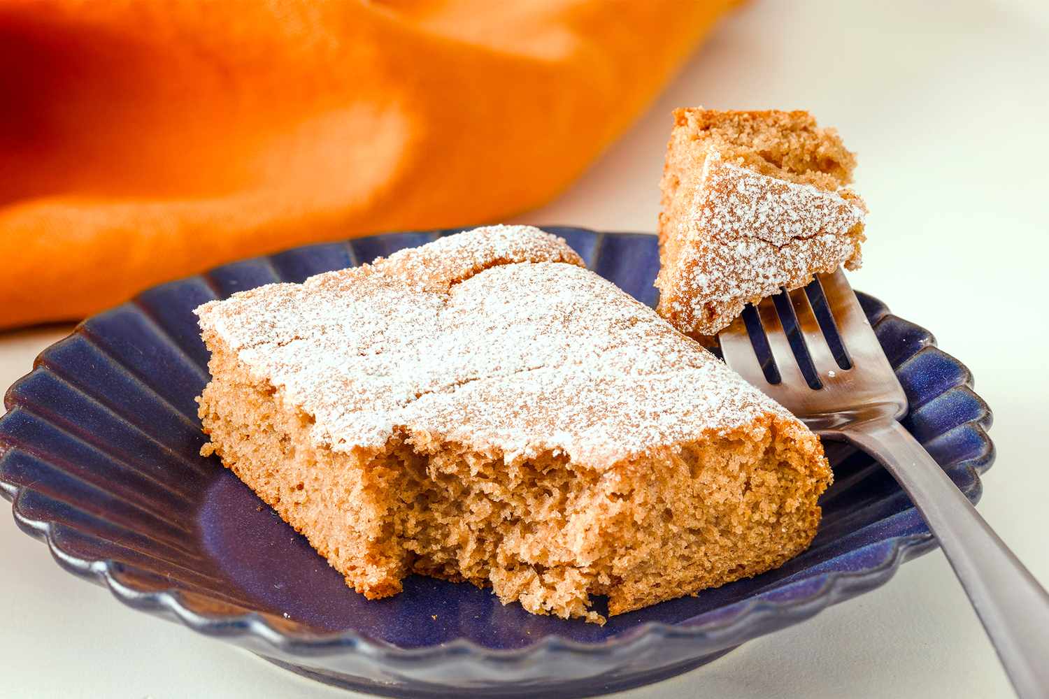 A slice of spice cake with powdered sugar on a plate with a fork cutting a piece
