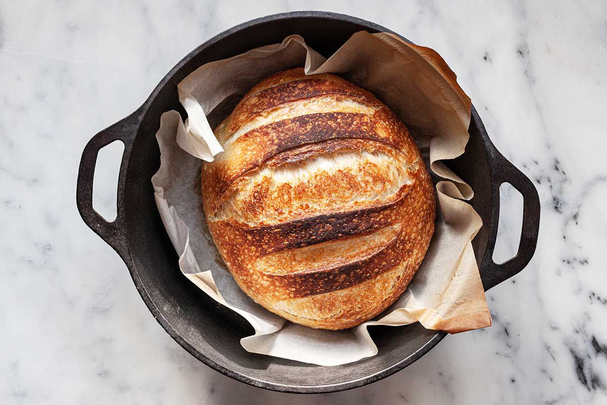 Showing How to Make No Knead Bread in a dutch oven. The loaf is golden and crusty inside the pot and has browned parchment paper underneath the loaf..