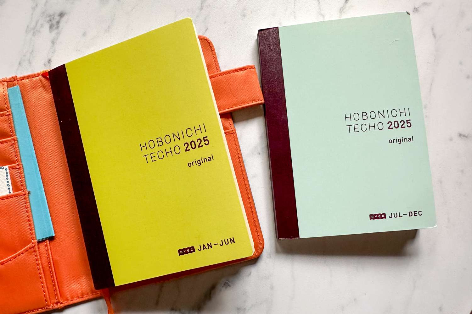 Two Hobonichi Techo 2025 planners one labeled JanJun and the other JulDec placed on a surface with an orange cover nearby