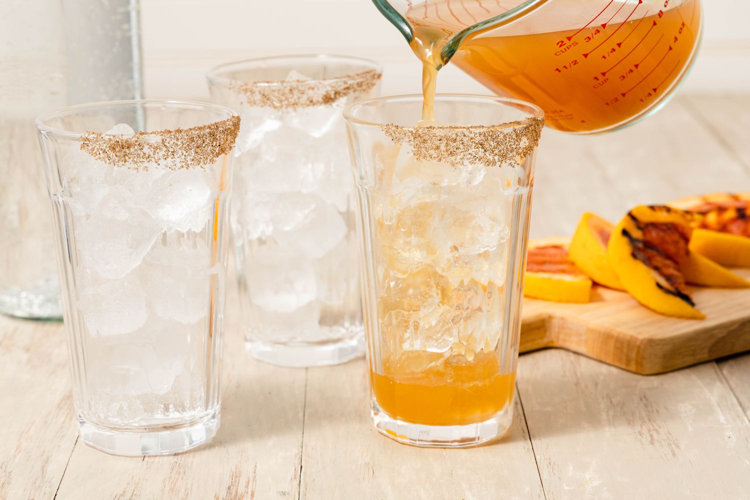 Grilled Grapefruit Paloma Poured Into a Salt Rimmed Glass With Ice, and in the Surroundings, Two More Salt Rimmed Glasses and a Cutting Board With Grapefruit Slices
