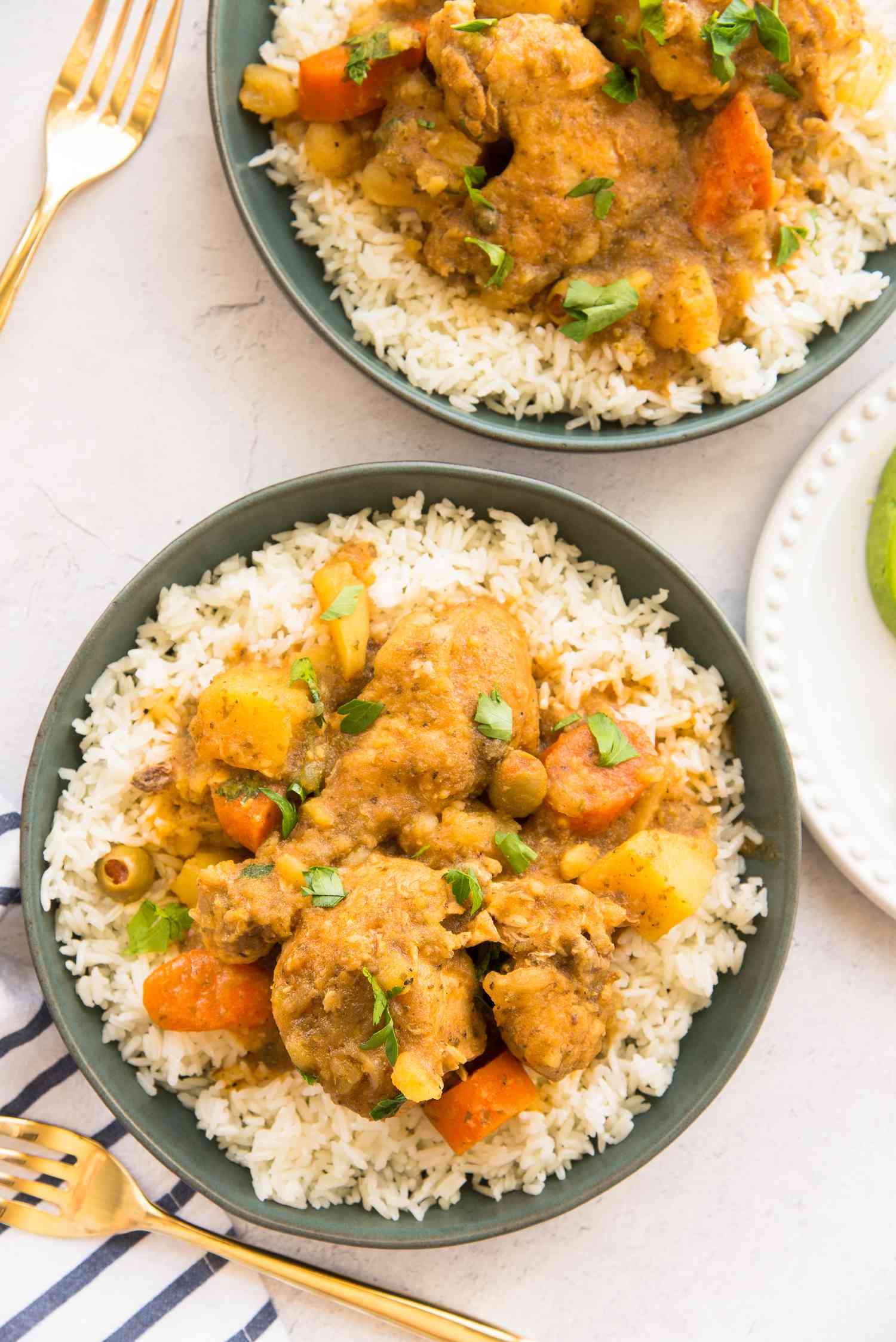 Two Bowls of Pollo Guisado Served Over Rice Next to Utensils 