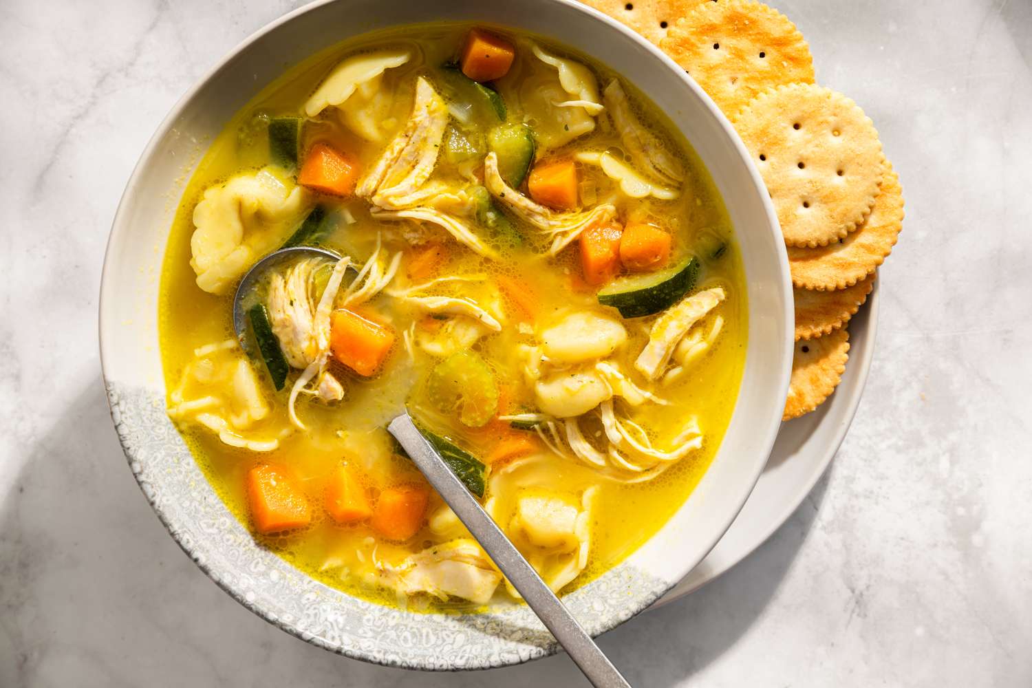 A bowl of chicken tortellini soup with vegetables served alongside crackers on a plate