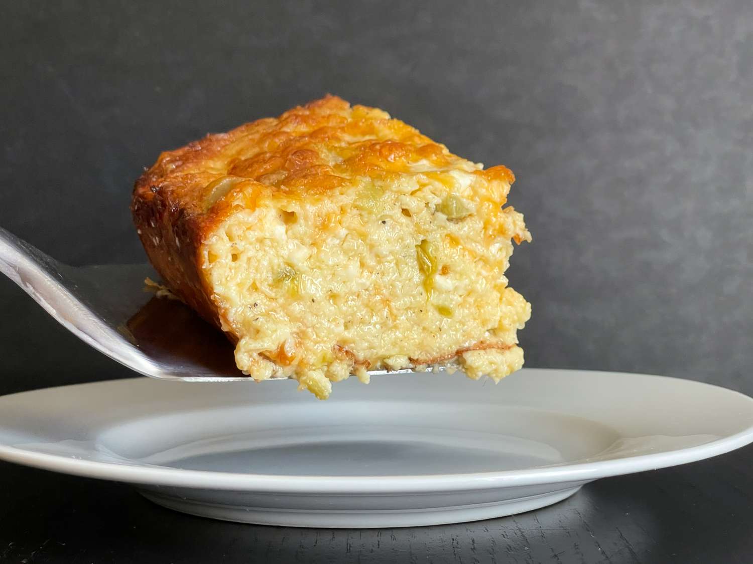 Side view of a slice of Chile Cheese Egg Casserole on a metal spatula held above a white plate