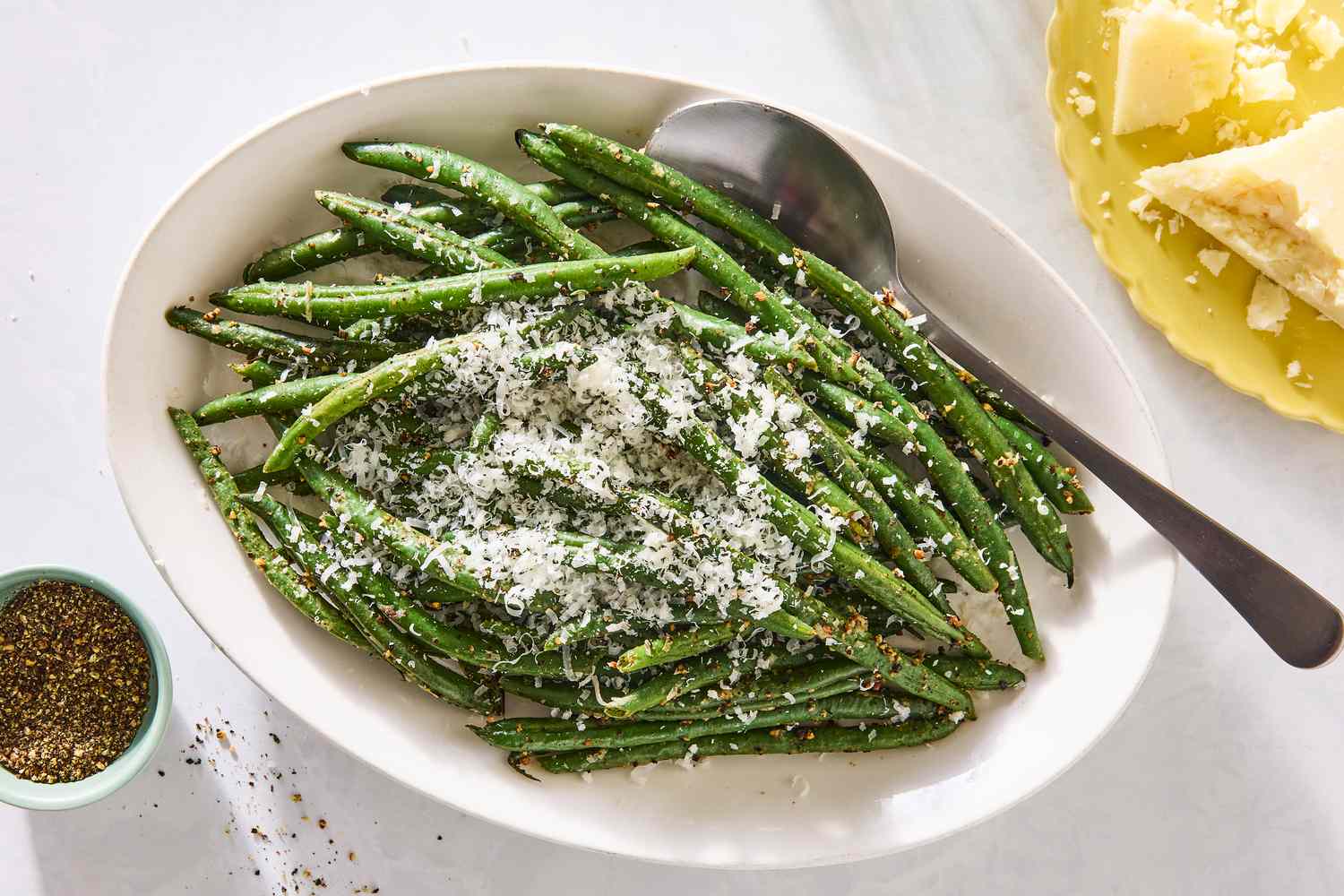 A dish of green beans topped with finely grated cheese served on a white oval plate with a spoon