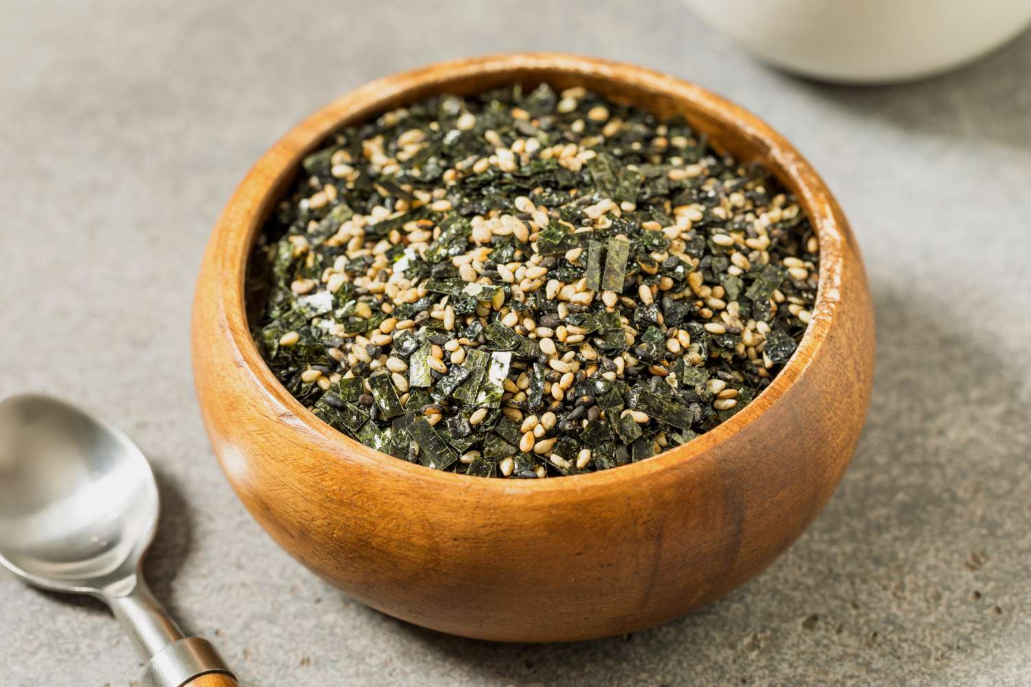 A wooden bowl filled with furikake a Japanese seasoning mix of nori seaweed and sesame seeds placed on a gray surface with a spoon nearby