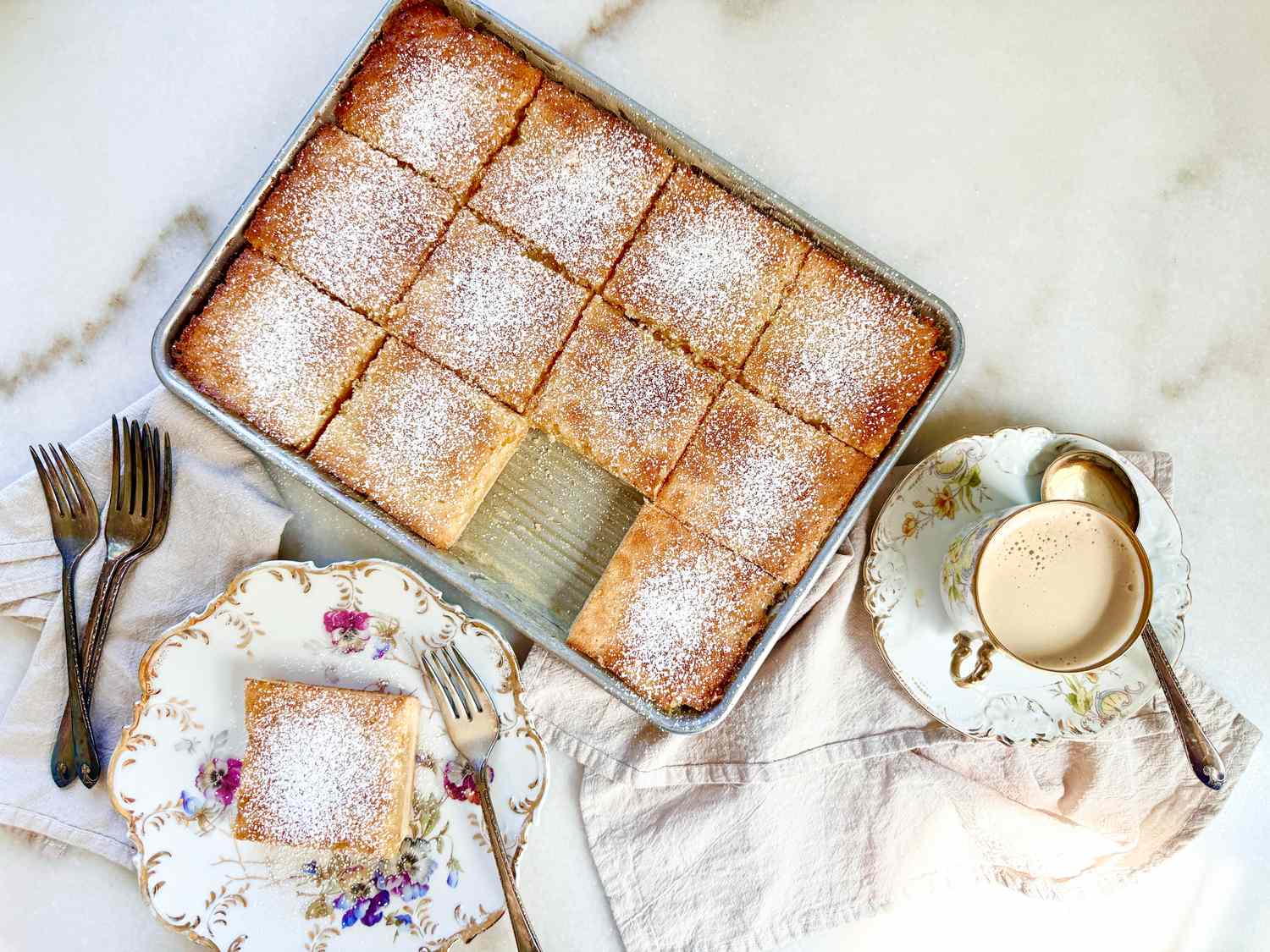 A tray of lemon squares dusted with powdered sugar, served with plates and forks, accompanied by a tea cup