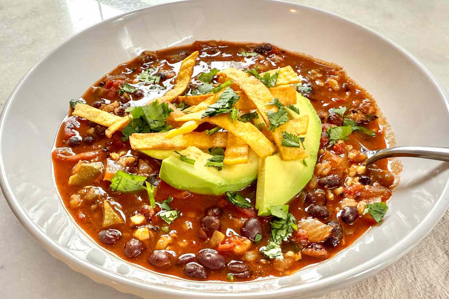 Angle view of a white bowl of Chrissy Teigen’s Veggie Tortilla Stew topped with tortilla strips, avocado and cilantro on a white countertop