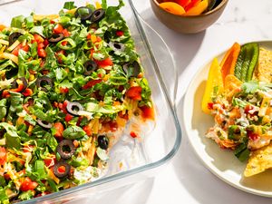 A glass dish containing layered nacho dip topped with ingredients such as olives herbs and vegetables served with tortilla chips on a plate