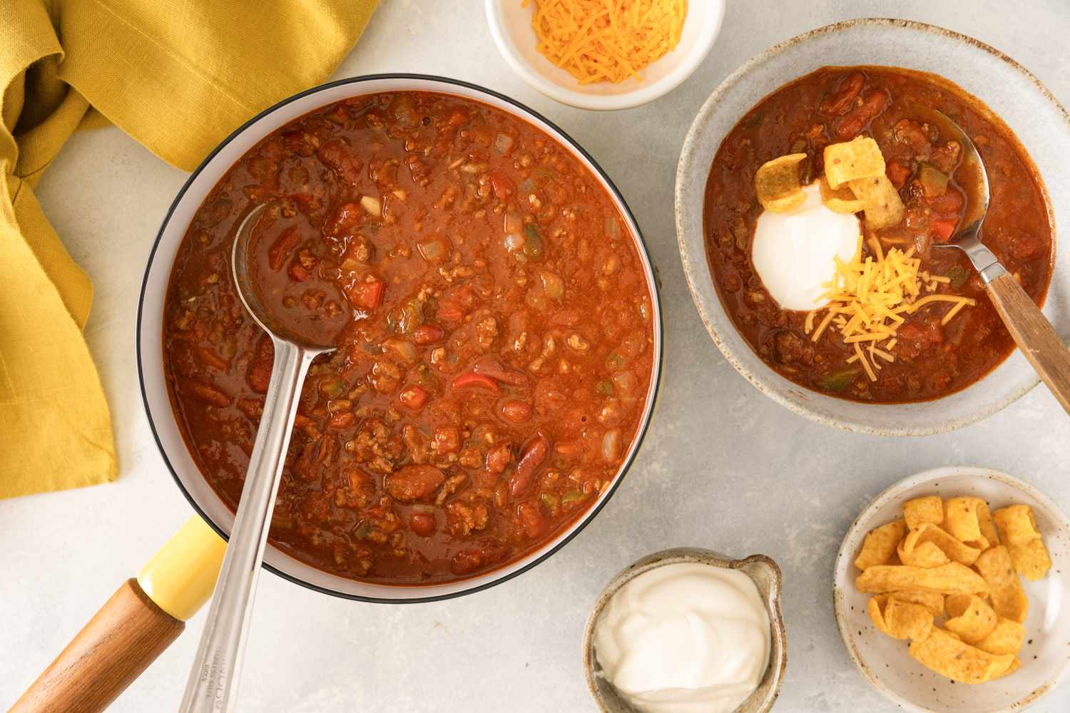 A pot of Grandma Patty’s Chili with a ladle next to a prepared bowl garnished with toppings
