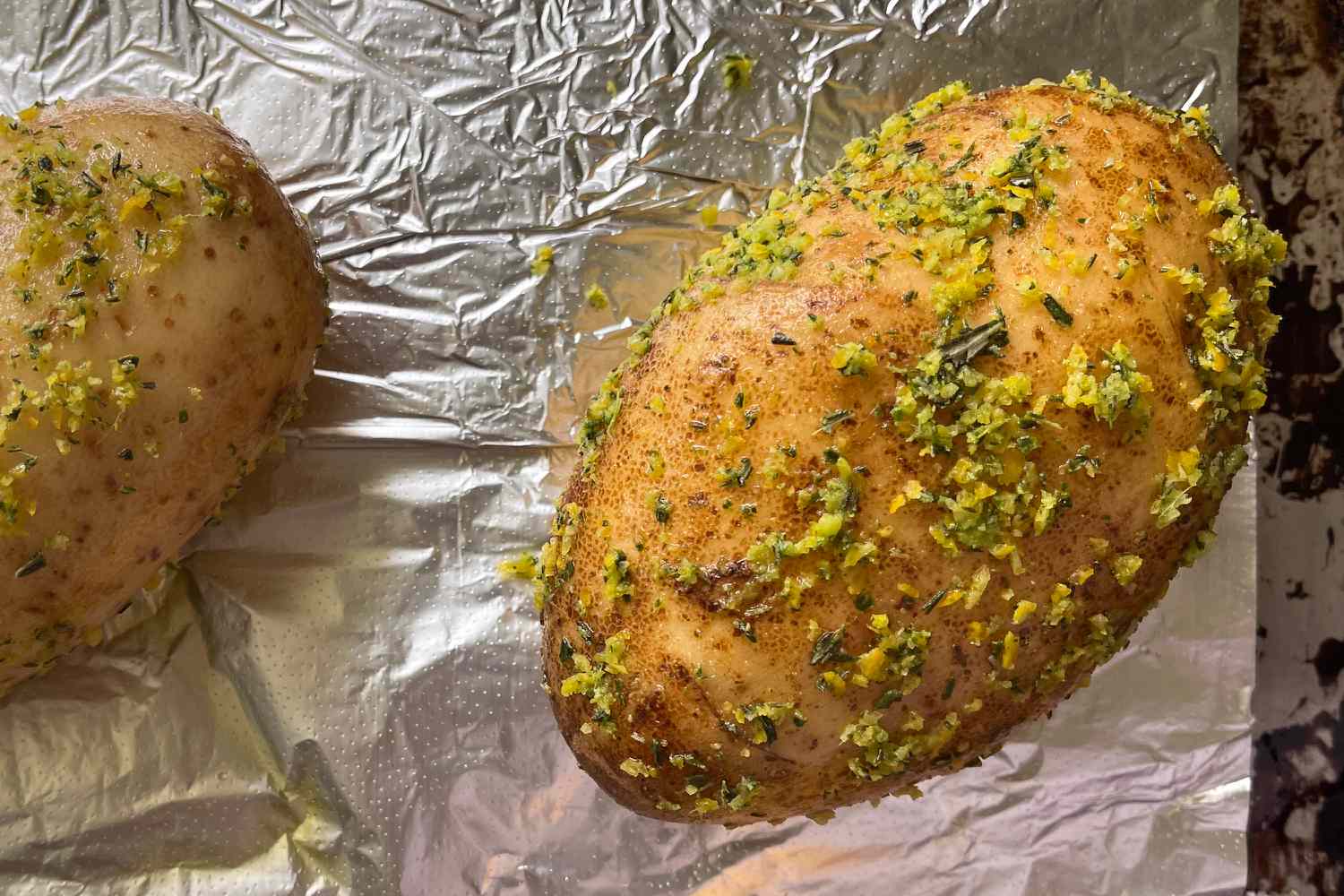 Overhead view of two baked potatoes with rub applied sitting on aluminum foil in a baking sheet