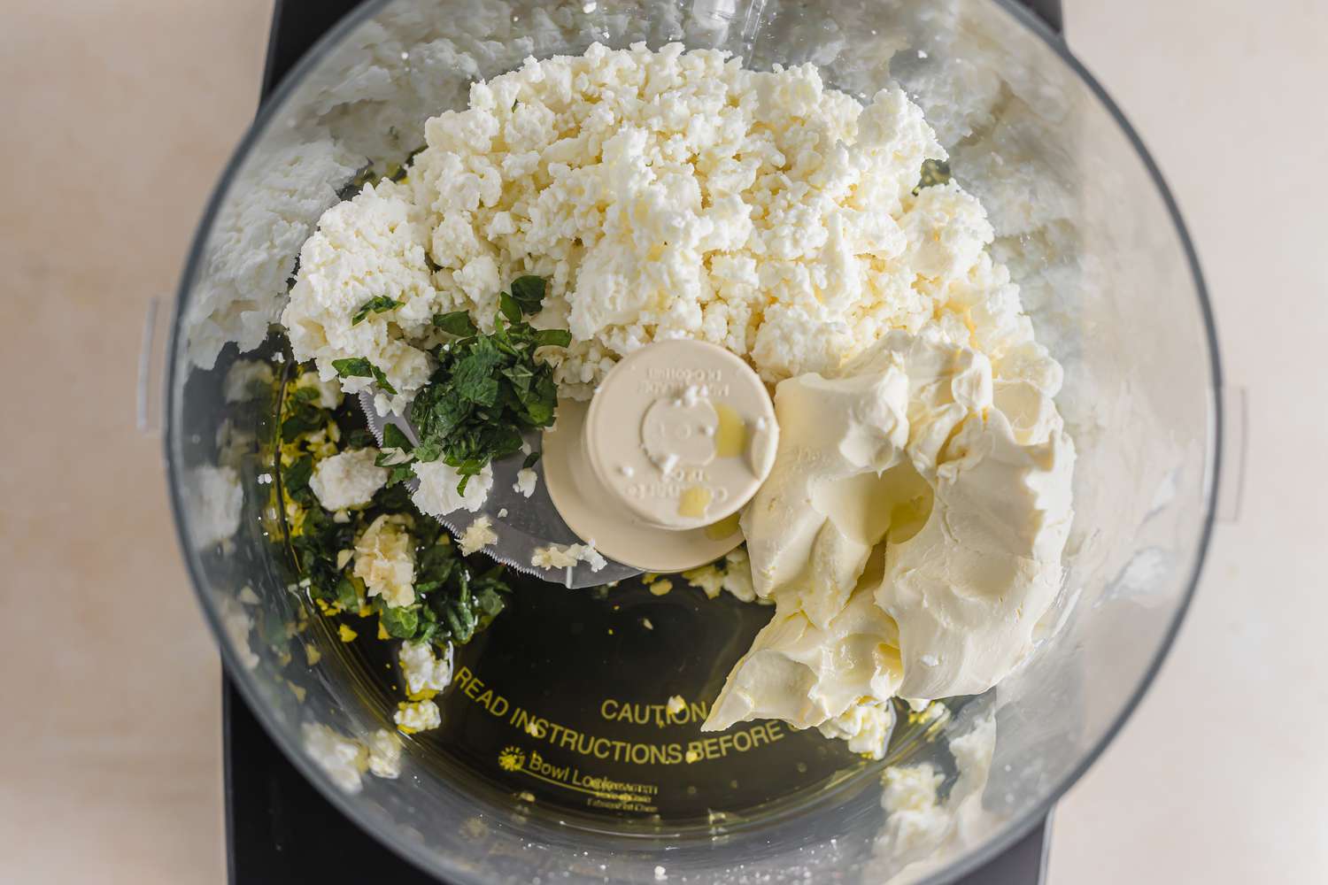 A food processor bowl with crumbled feta and other ingredients to make whipped feta with figs, pistachios, and a honey drizzle