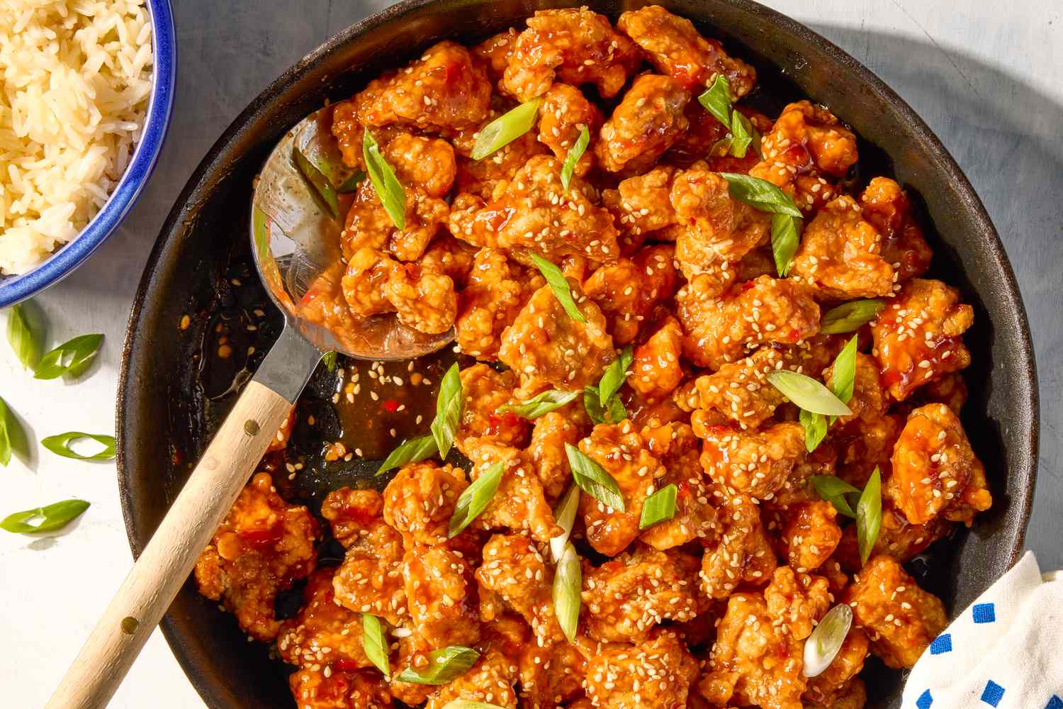 General Tsos chicken dish with sesame seeds and green garnish in a skillet served with rice on the side in a bluerimmed bowl