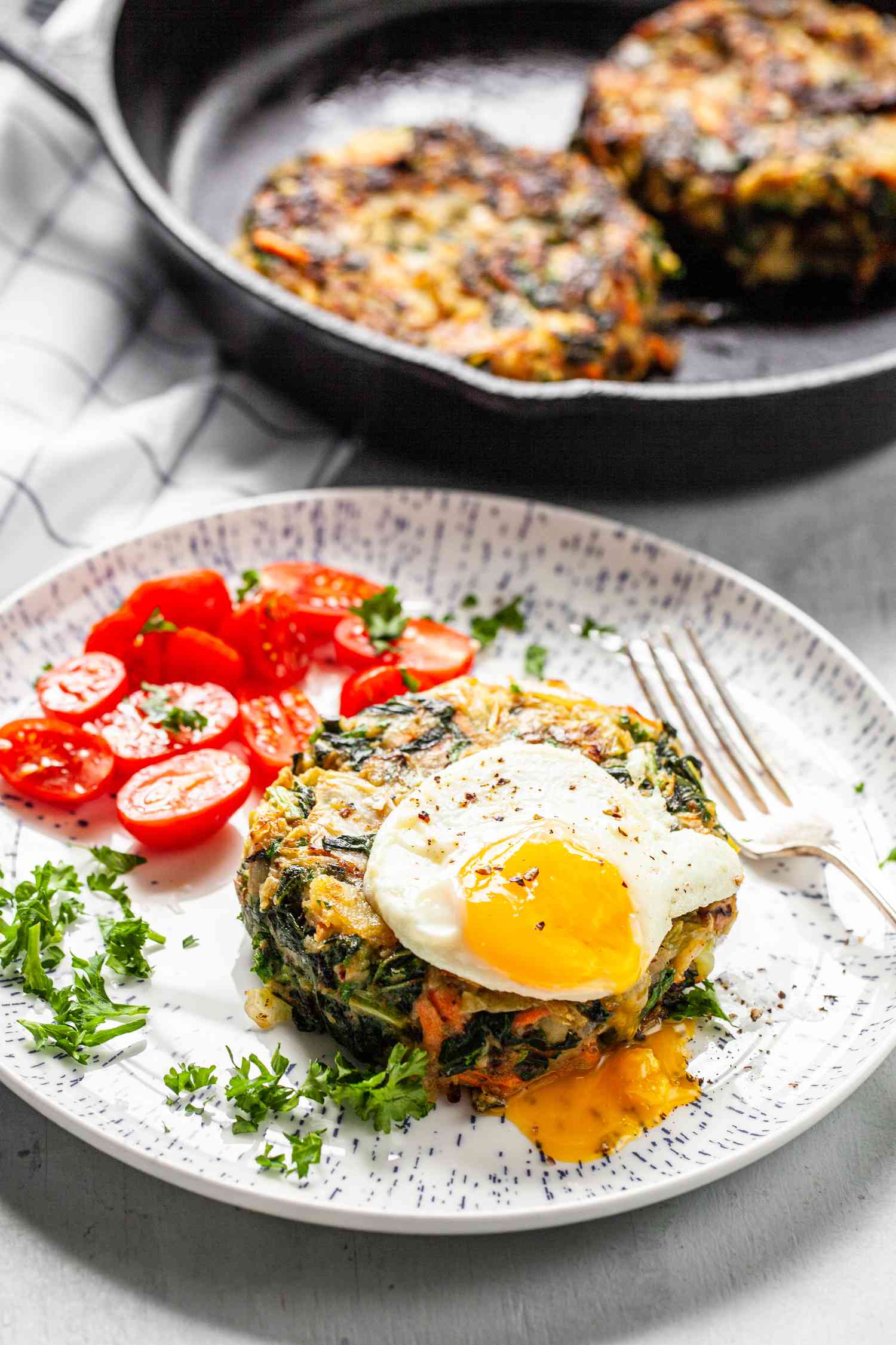 Bubble and Squeak Topped with Sunny Side Egg on a Plate with Halved Cherry Tomatoes and a Fork, Next to a Cast Iron Skillet with More Patties