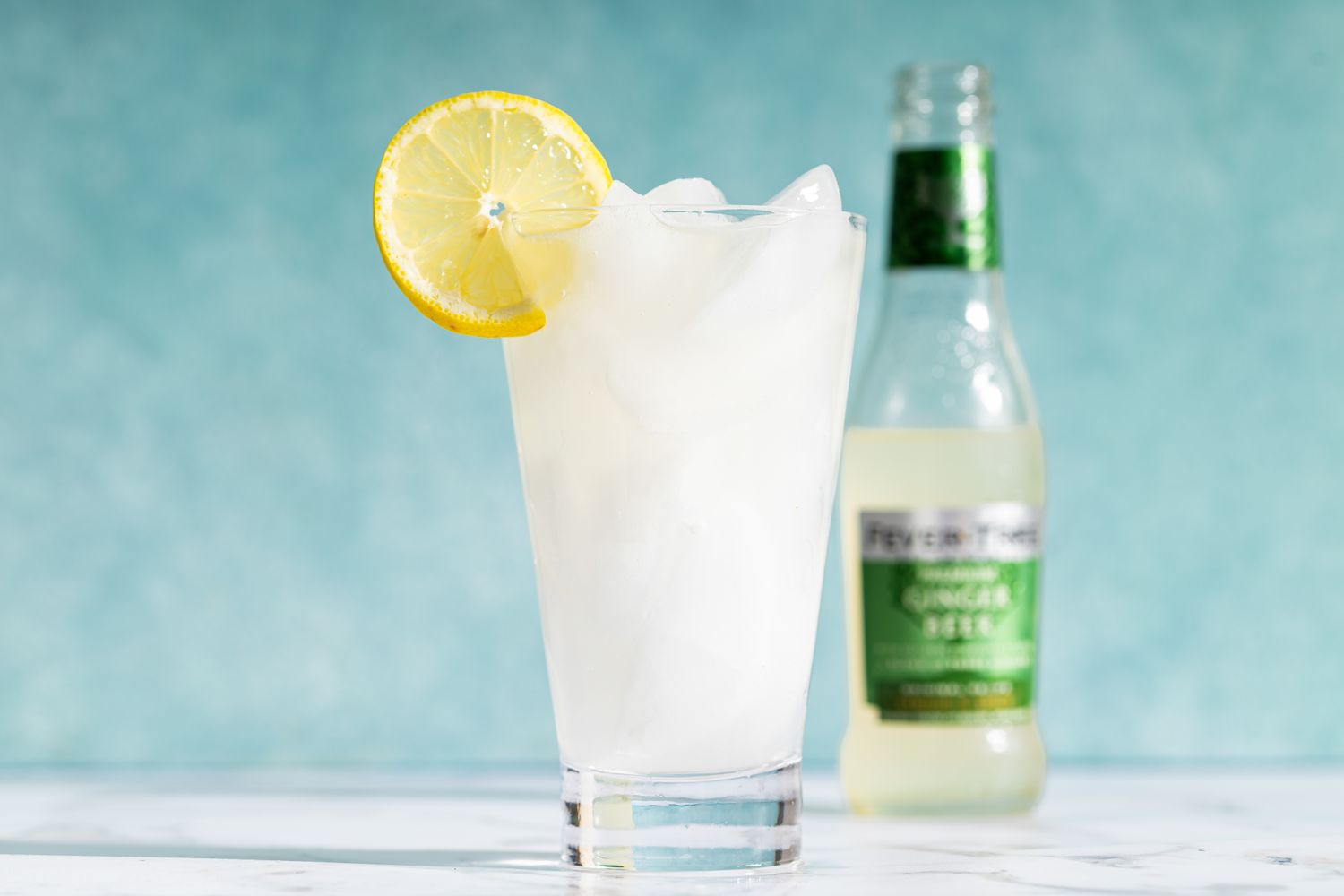 Lemonade glass with ice and lemon slice ginger ale bottle in background