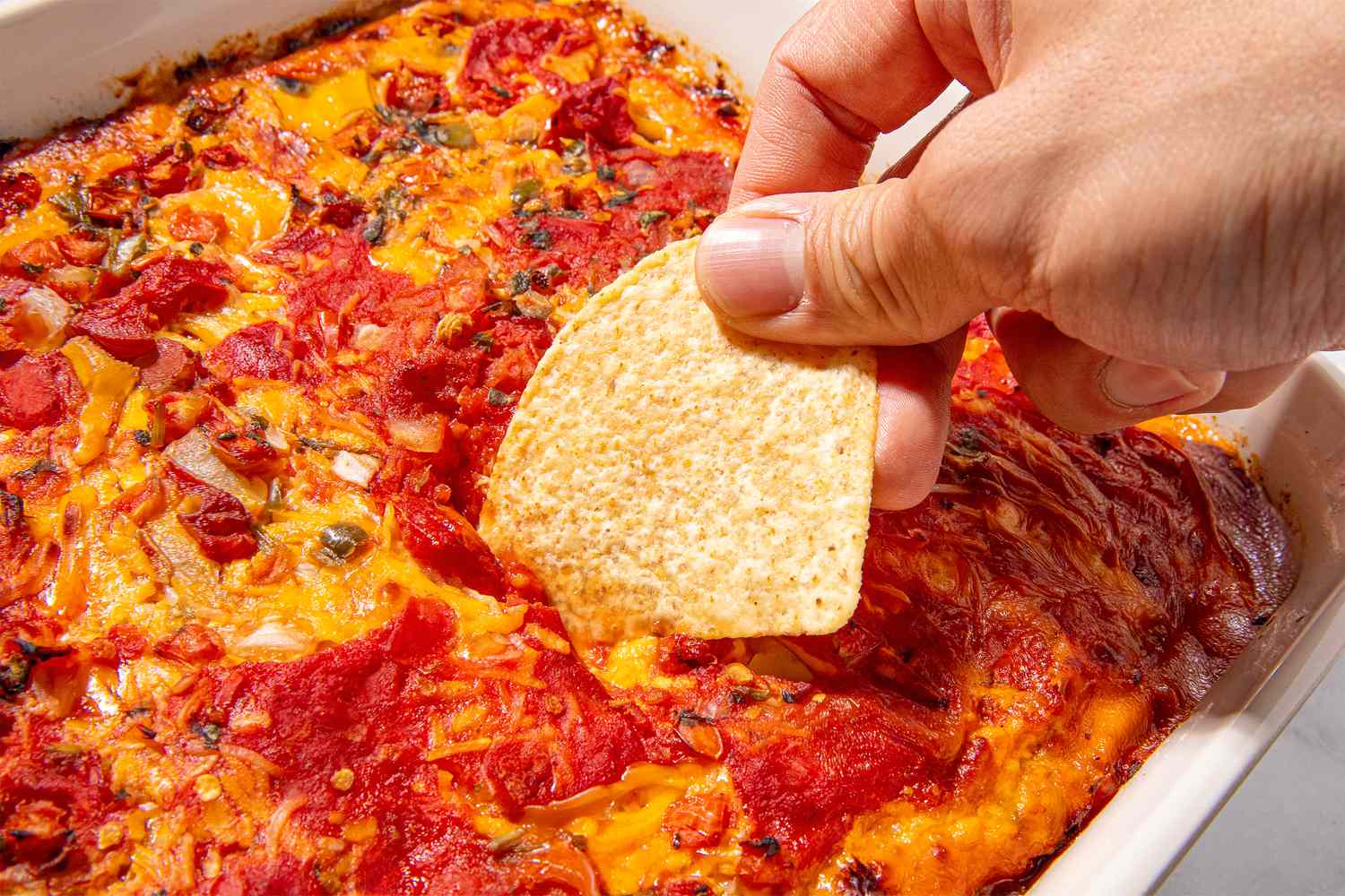Hand dipping a tortilla chip into a baked tomato and cheese dip