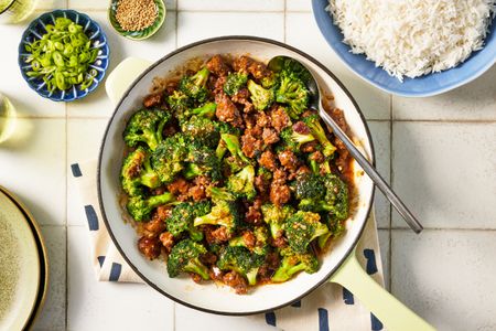 Bowl of ground beef and broccoli stir fry at a table setting with a bowl of rice, bowl of sesame seeds and a glass of water