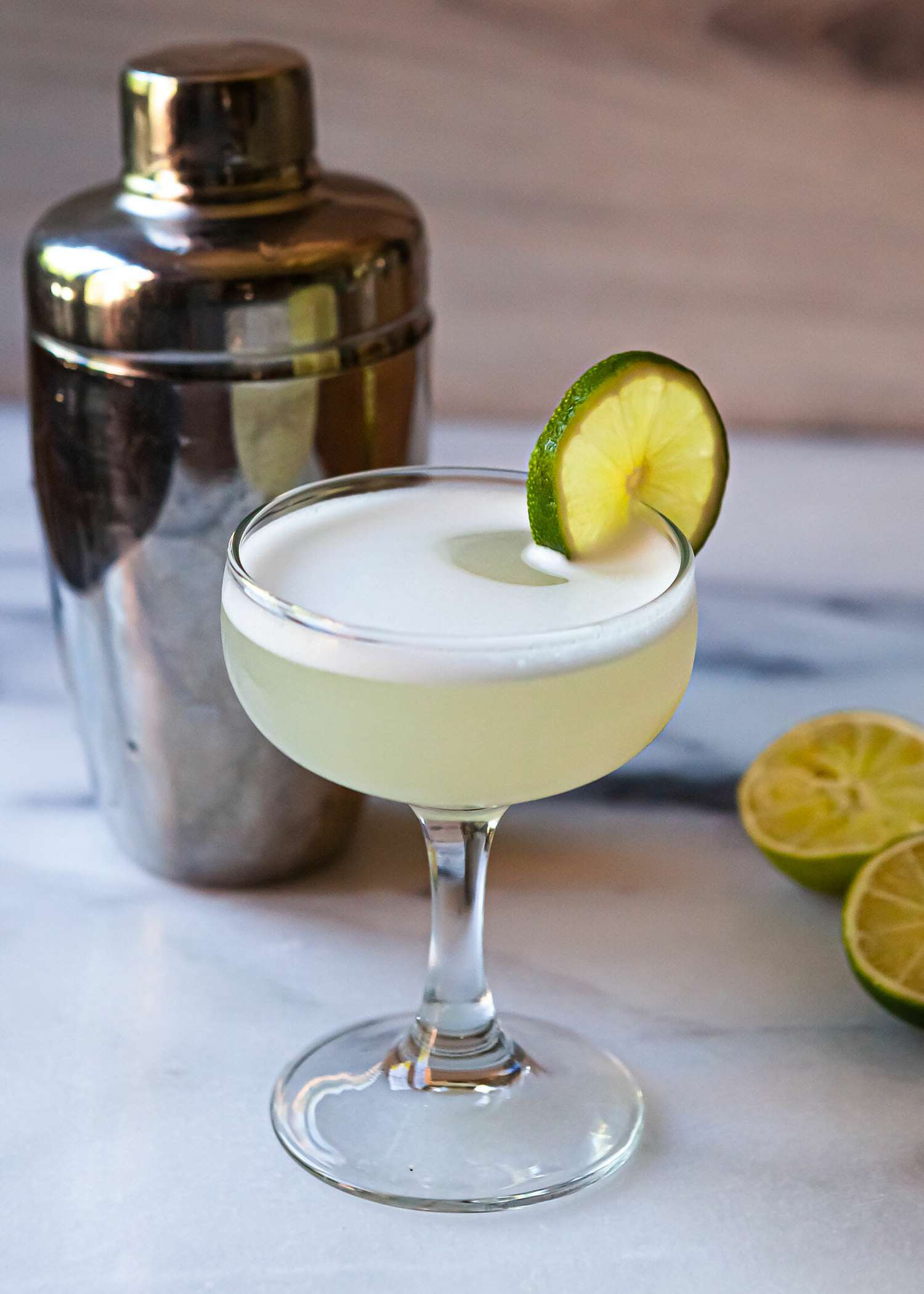 A gimlet recipe in a chilled coupe glass with a cocktail shaker behind it and limes to the right.