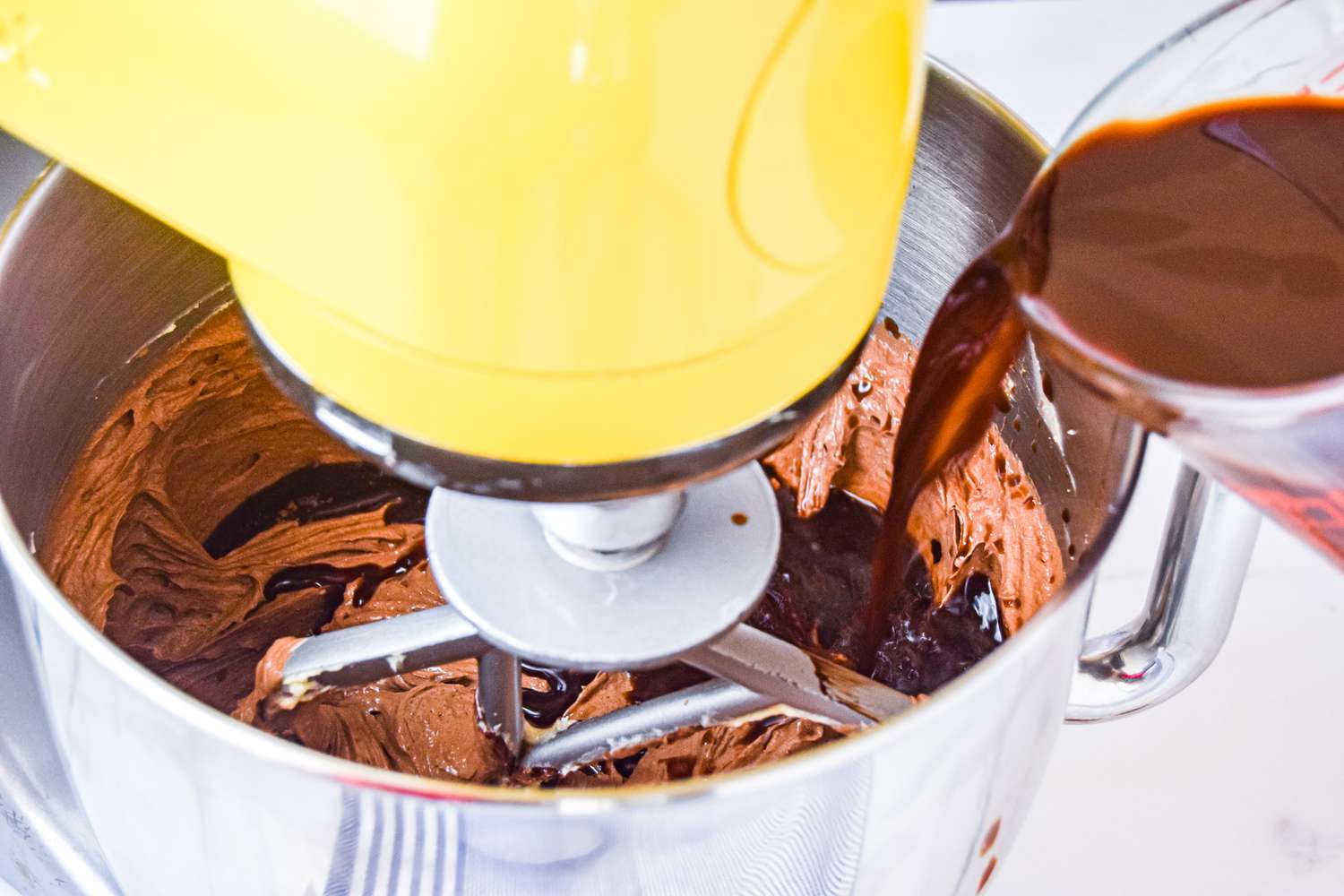 Chocolate Bourbon Cake batter in a mixing bowl with a measuring cup pouring in a coffee-bourbon mixture