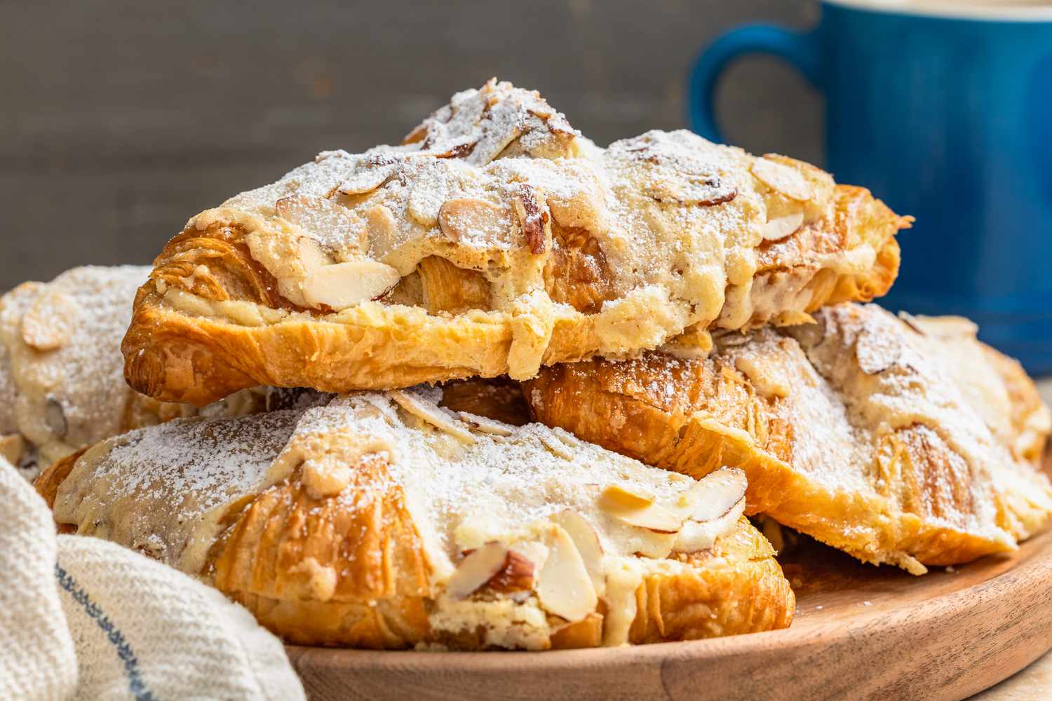 Pile of Almond Croissants on a Wooden Tray, and in the Surroundings, a Kitchen Towel and a Blue Coffee Mug