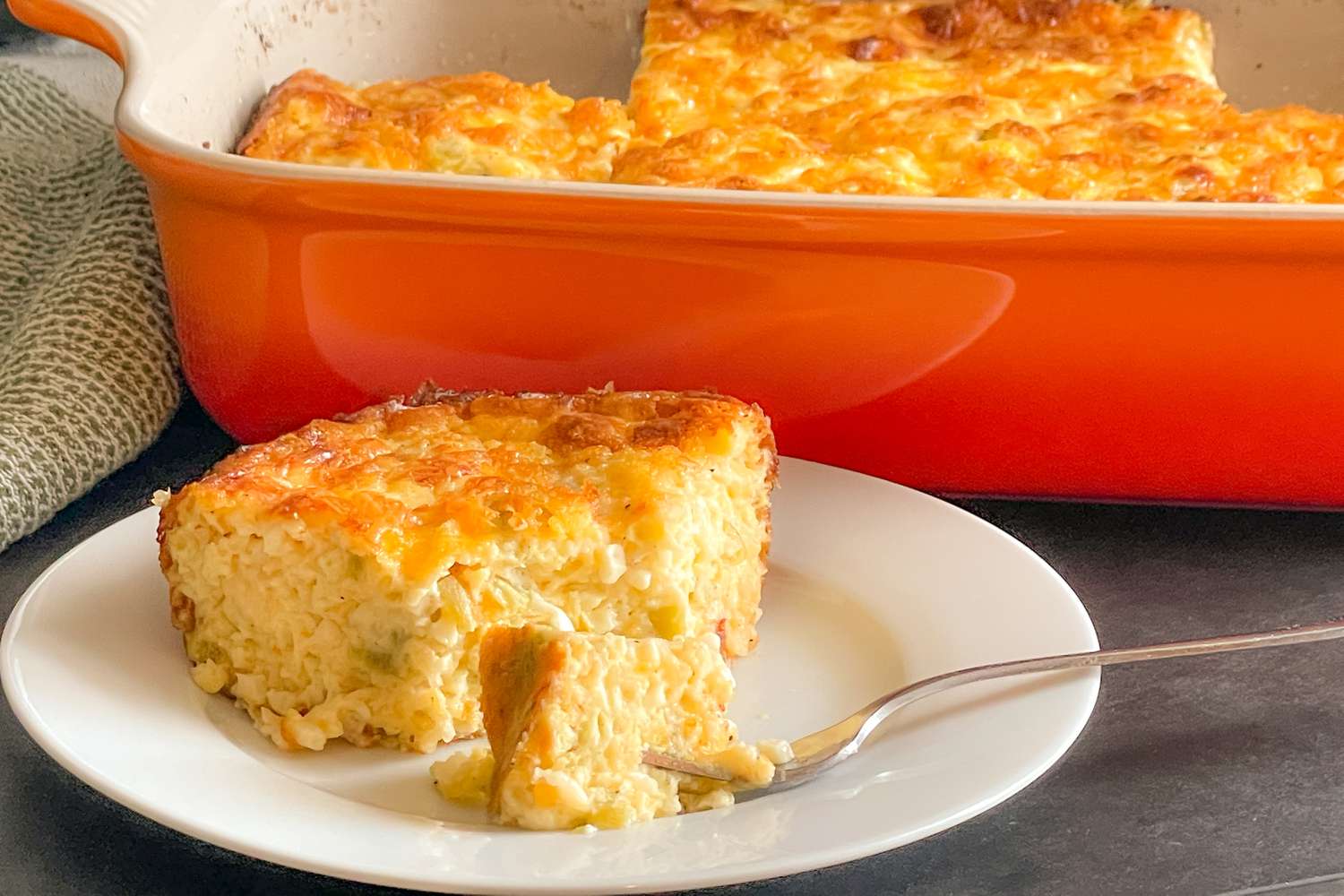 Angled view of a slice of Chile Cheese Egg Casserole on a white plate with a fork and the orange casserole dish in the background