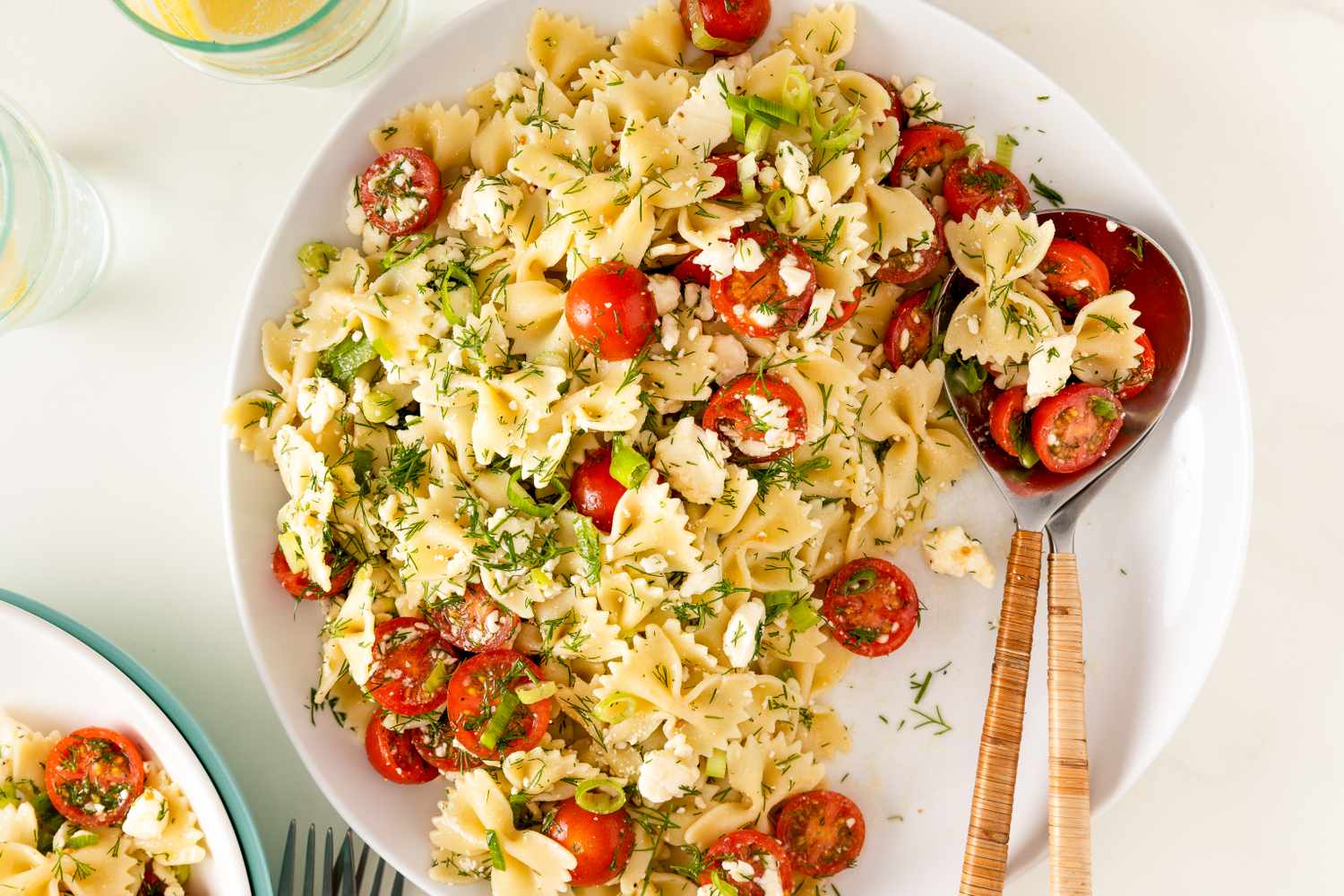 A plate of Greek pasta salad with tomatoes, feta cheese, and herbs, served with a wooden-handled serving spoon