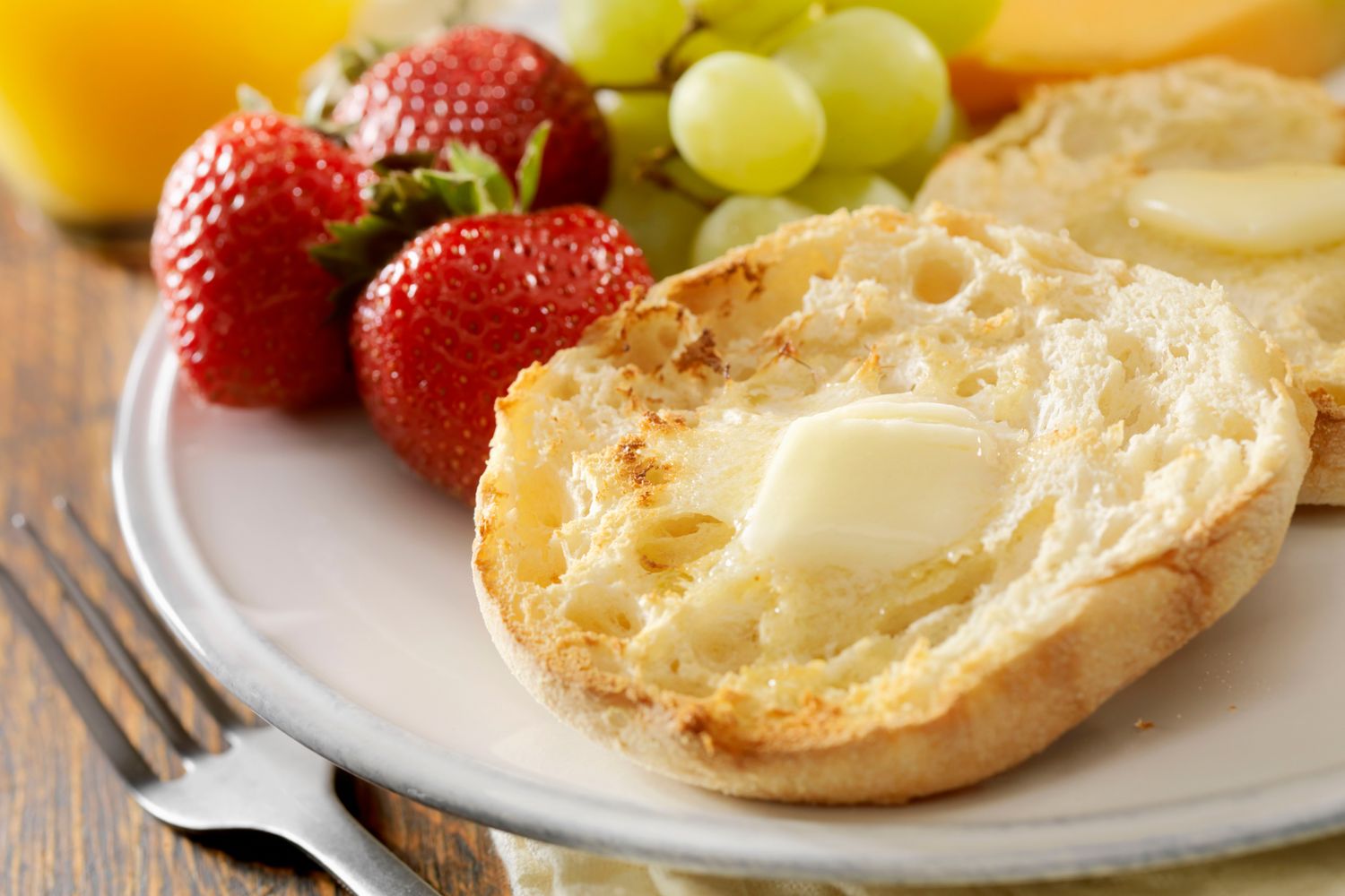 English muffin topped with butter on a plate accompanied by strawberries and grapes