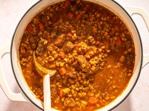 A pot of beef barley soup with visible chunks of meat and vegetables and a wooden spoon
