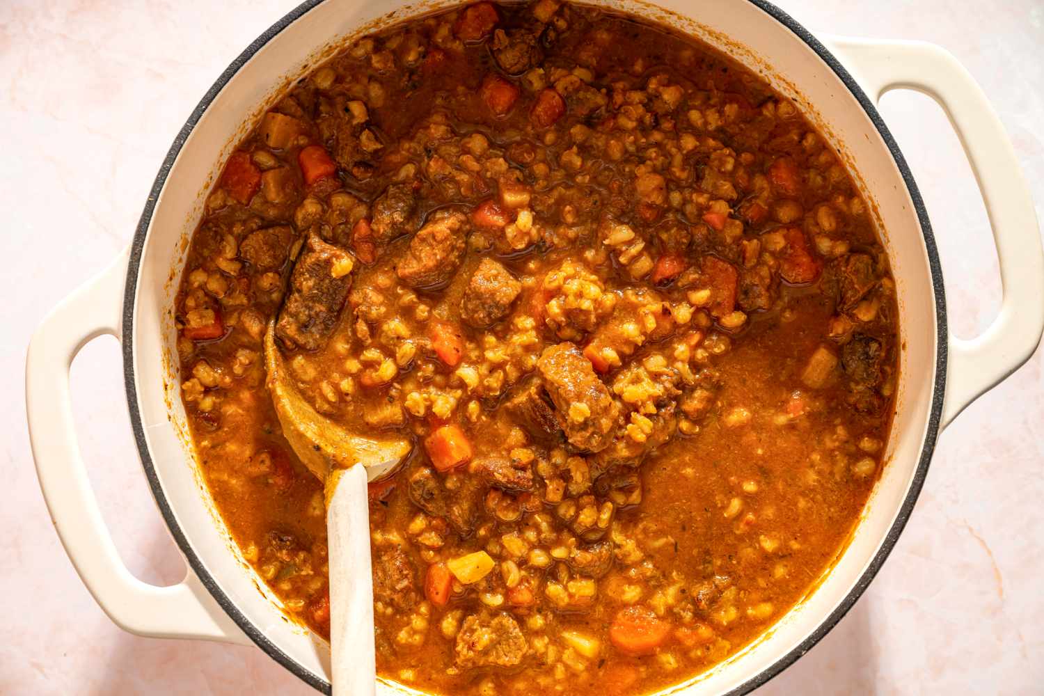 A pot of beef barley soup with visible chunks of meat and vegetables and a wooden spoon