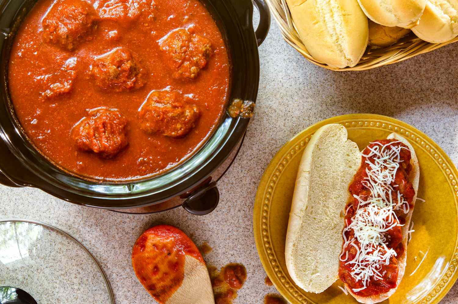 Slow cooker with meatballs in marinara sauce a toasted sandwich with meatballs and cheese on a yellow plate and a basket of rolls