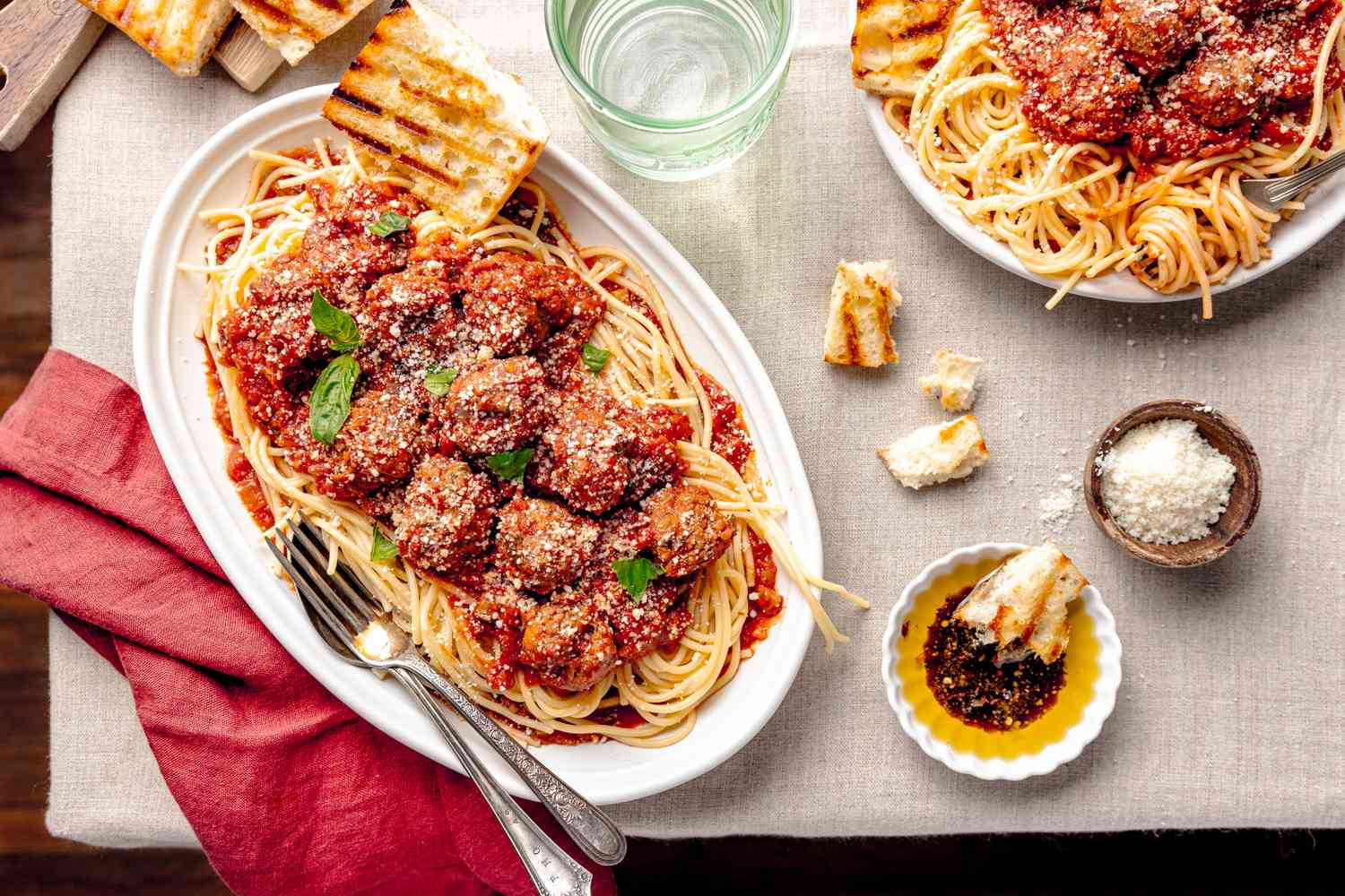 Table Setting (L to R): Toasted Bread on a Wooden Board, Burgundy Table Cloth, a Platter of Spaghetti and Meatballs With a Slice of Tasted Bread and Topped With Basil, a Glass of Water, Bite Size Bread Pieces on the Table, a Bread Piece Dipped into a Small Saucer With Olive Oil and Crushed Red Peppers, a Small Bowl With Parmesan, and a Plate With More Spaghetti and Meatballs