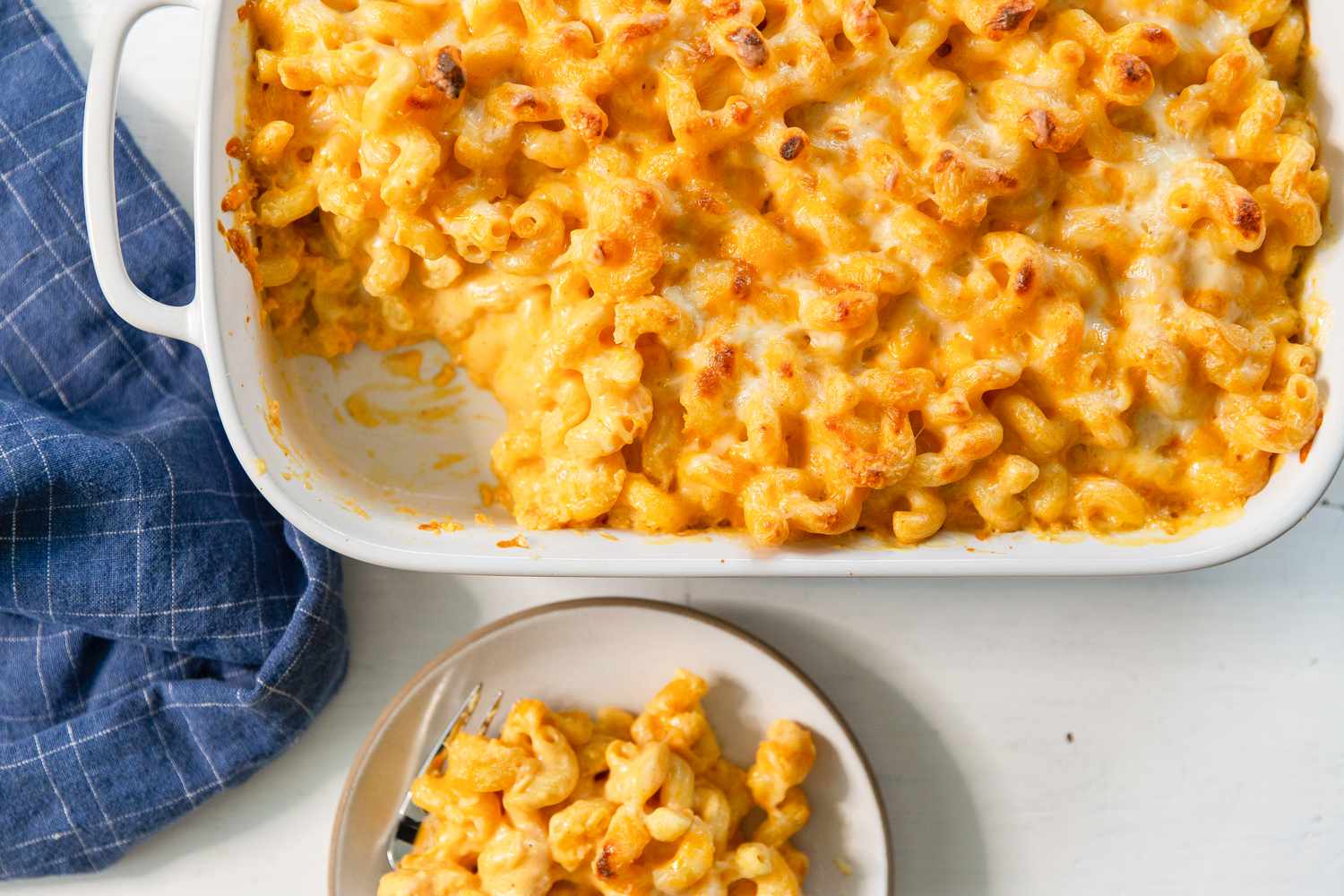 Overhead view of a white baking dish and small plate of macaroni and cheese next to a blue table napkin