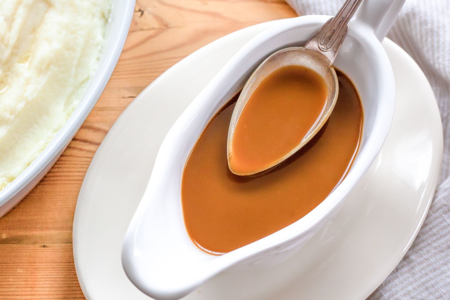 Overhead view of a gravy boat of gravy and a spoon full of gravy next to a plate of mashed potatoes and a white napkin all on a wooden tabletop