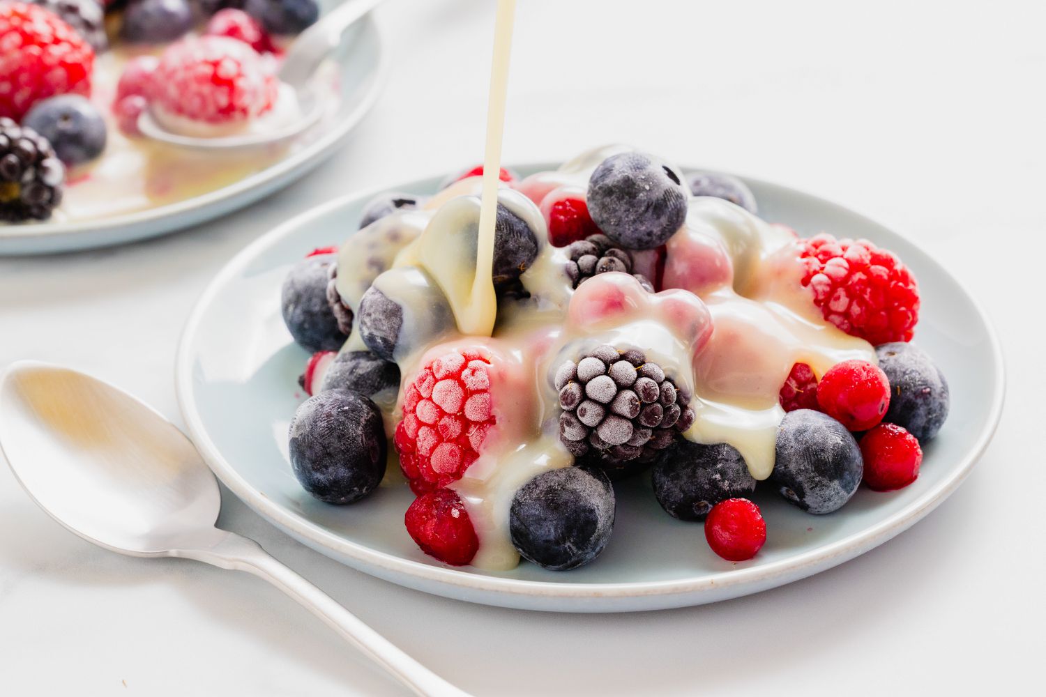 A plate of mixed berries topped with white chocolate sauce being poured, served with a spoon on the side