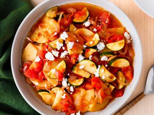Overhead view of a white bowl of Zucchini Con Patate on a table next to a spoon and a green napkin