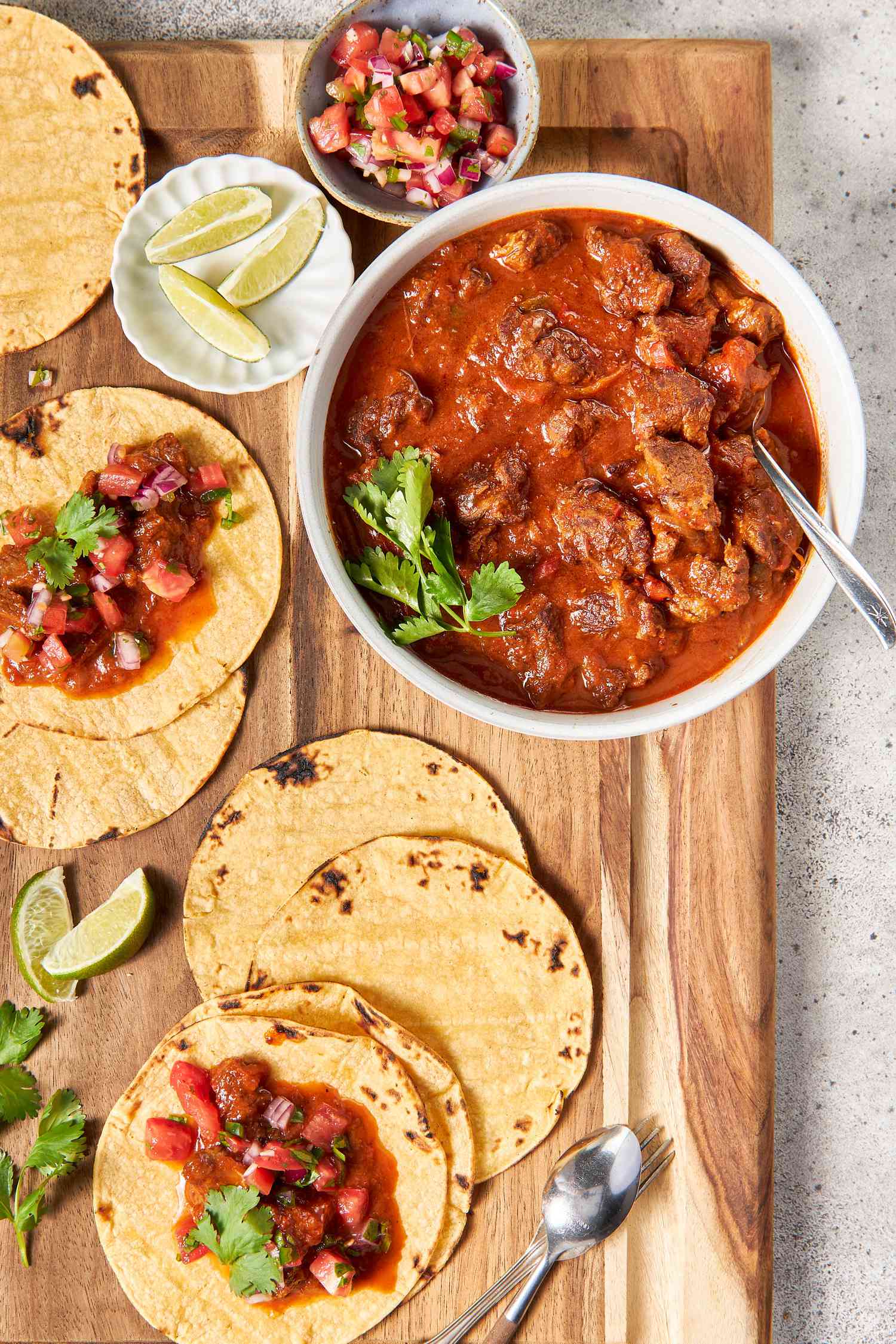 On a wooden board, a bowl of carne picada with a sprig of cilantro and a spoon, surrounded by a bowl of pico de gallo, a small plate of lime wedges, and tortillas spread all over the board (some with carne picada and pico de gallo, some plain)