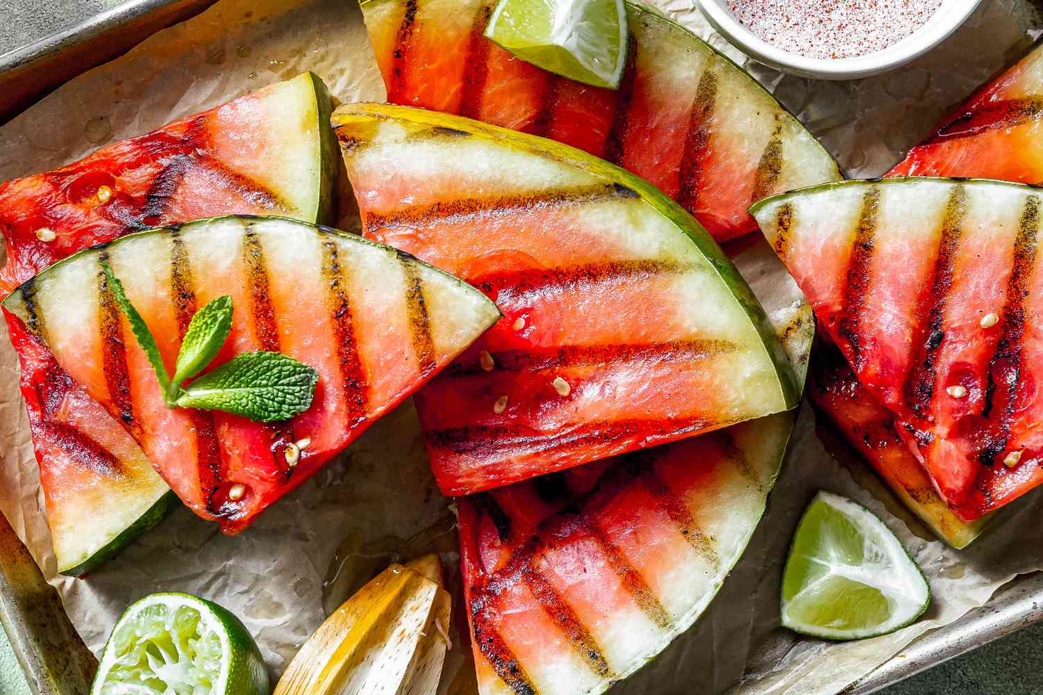 Grilled Watermelon Slices Topped With Mint Sprigs and Lime Wedges and Next to a Bowl of Spice Blend, All on a Parchment Paper Lined Baking Tray