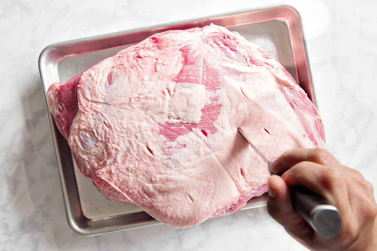 A baking sheet rests on a marble background with a raw pork shoulder in it. A hand with a chef's knife is cutting deep slits in the roast.