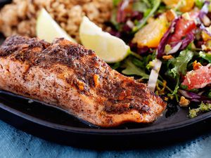 Side view of a fillet of broiled jerk salmon. The salmon has juices pooled at the bottom of it. A mixed green salad with citrus, two lime wedges and rice are visible on the plate behind the salmon. The plate is on a blue linen.