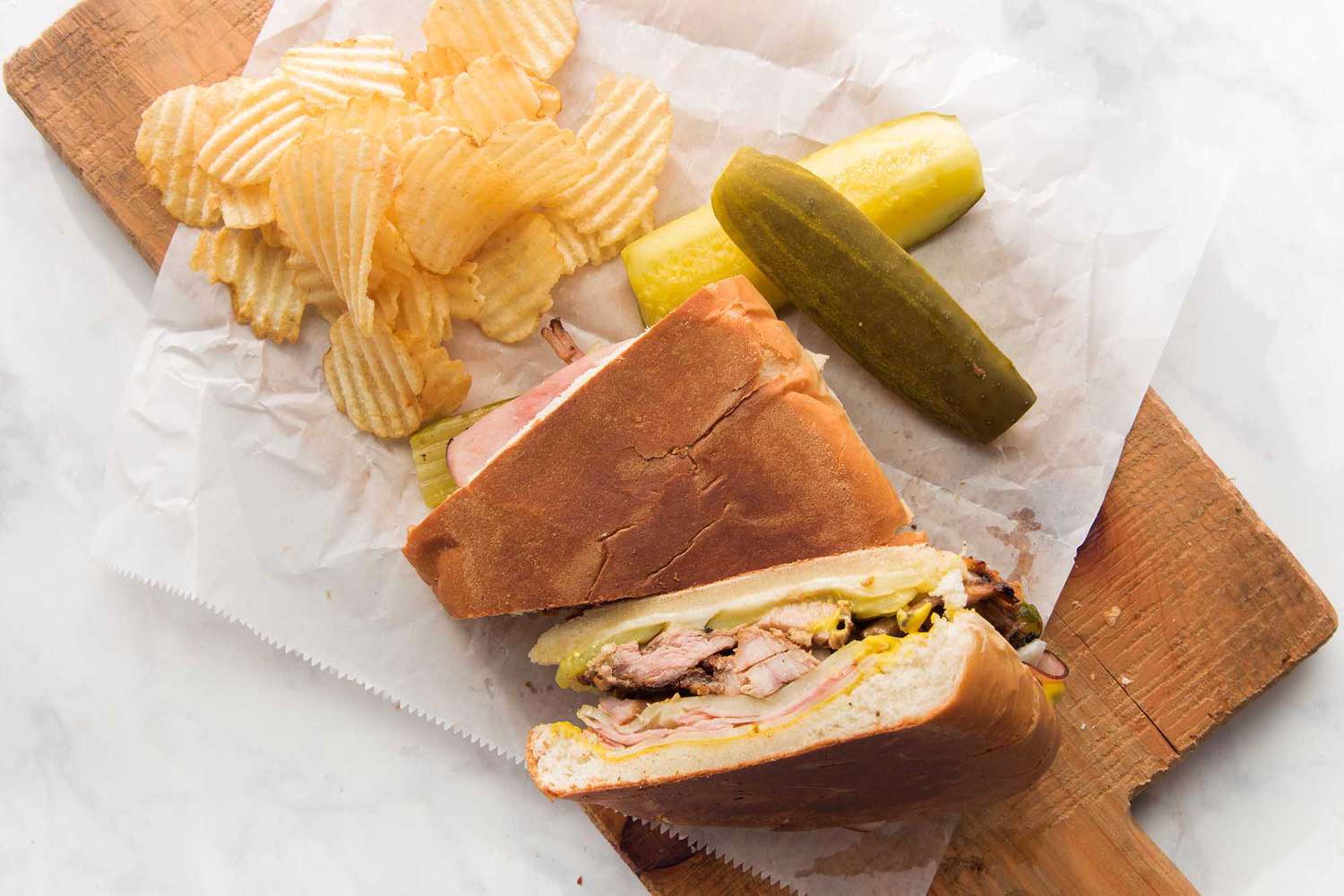 A rectangular cutting board is set on a marble background. Two halves of the best cuban sandwich are set on top, along with ridged potato chips and pickle spears
