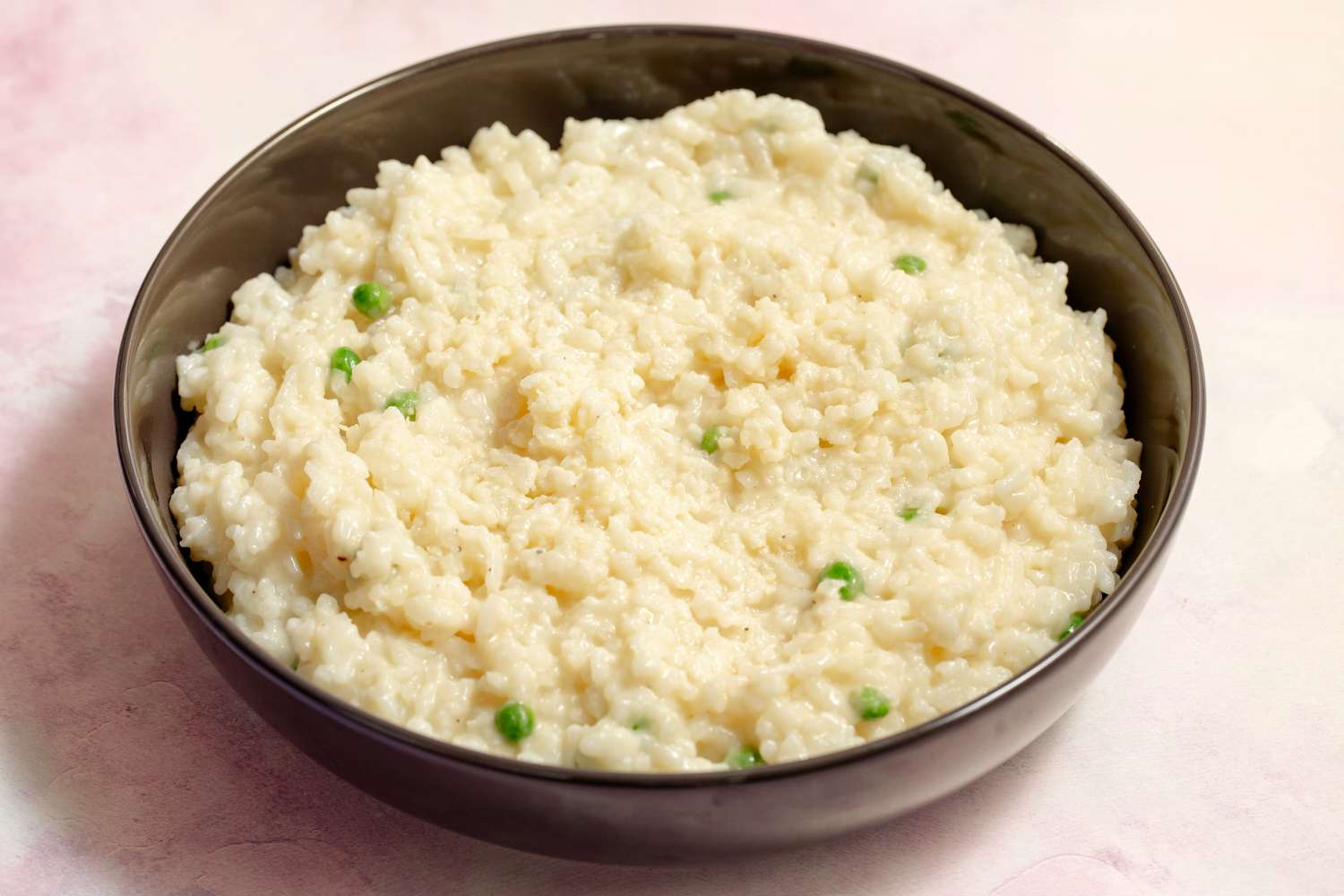 Closeup view of a brown bowl of risotto with peas on a pink tinted countertop