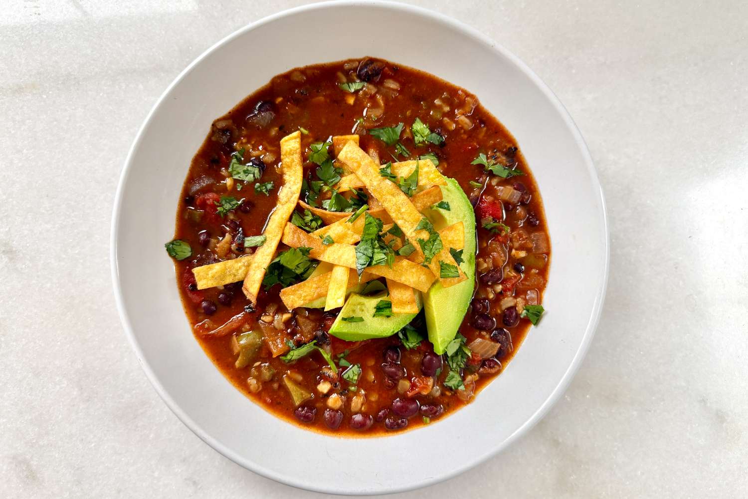 Overhead view of a white bowl of Chrissy Teigen’s Veggie Tortilla Stew topped with tortilla strips, avocado and cilantro on a white countertop
