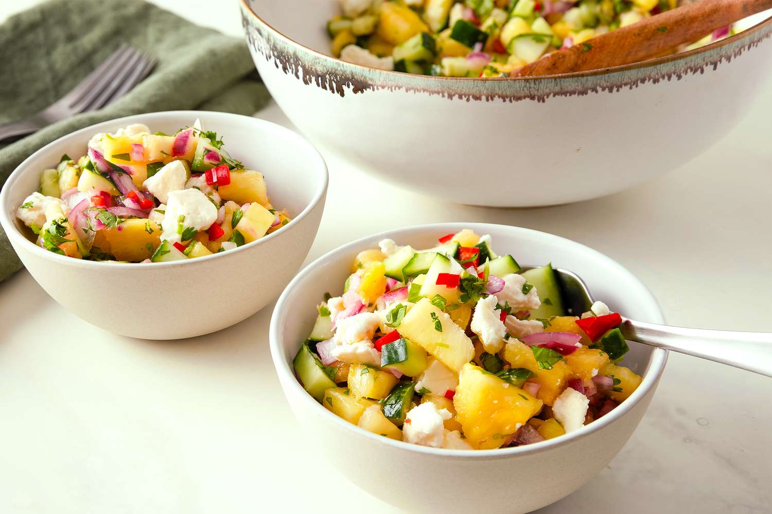 2 small bowls of Pineapple Cucumber Salad with Feta with forks and napkins and a large serving bowl of the salad with a wooden spoon