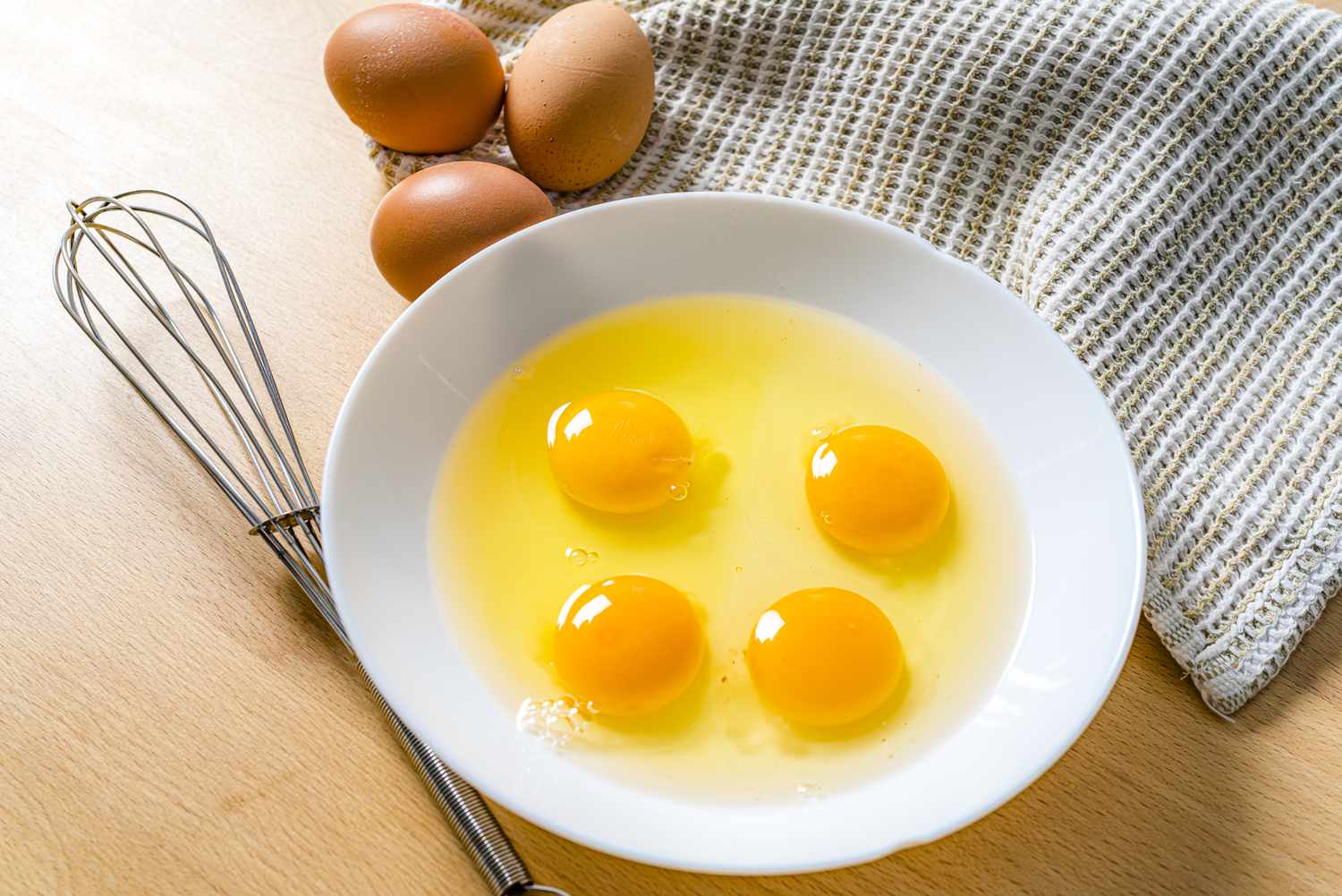 Four cracked raw eggs in a bowl next to a whisk and three whole eggs