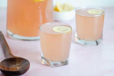 Two glasses of pink lemonade garnished with lemon rounds. A wooden spoon and pitcher are next to the glasses.