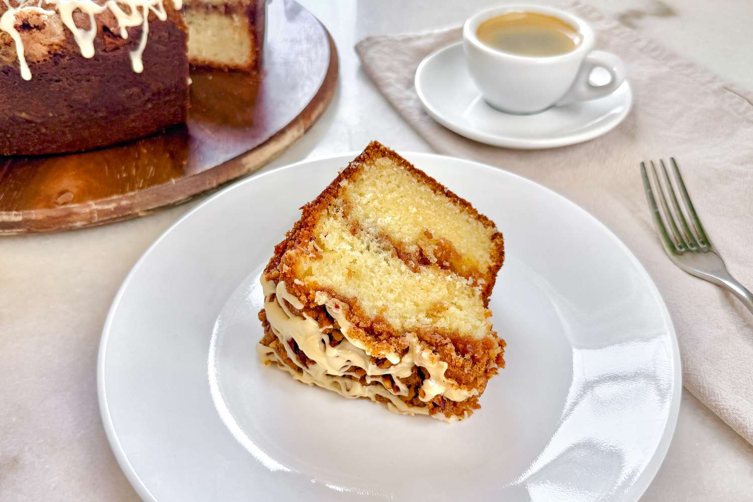 A slice of coffee crumble cake on a white plate next to a cup of coffee on a table