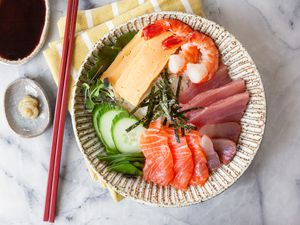 Chirashi Bowl with Salmon, Tuna, Shrimp, Tamagoyaki, and Sliced Cucumbers Next to a Small Saucer of Wasabi and Another Small Saucer of Soy Sauce