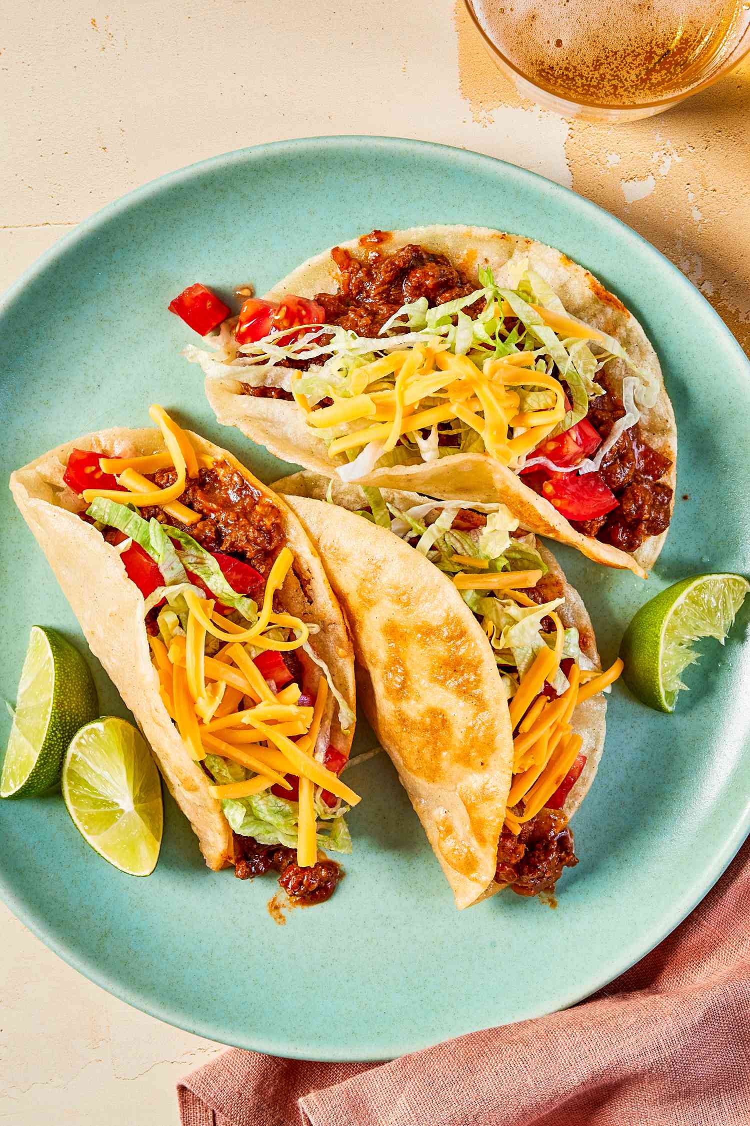Crispy ground beef tacos on a plate at a table setting with glasses of beer and a table napkin