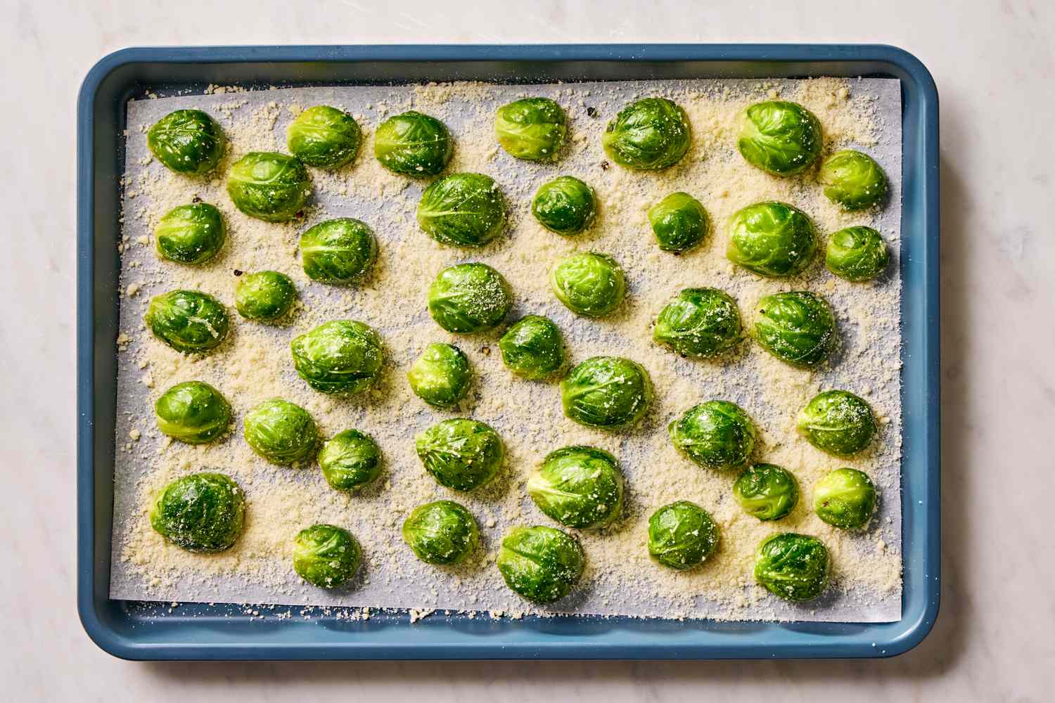 Overhead view of a blue baking sheet lined with parchment paper with brussels sprout slices and parmesan added for Crispy Parmesan Brussels Sprouts Recipe