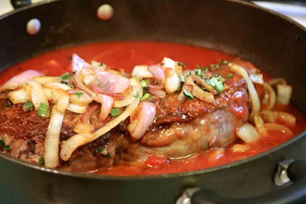 A steak cooking and covered in herbs, onions, and a tomato purée