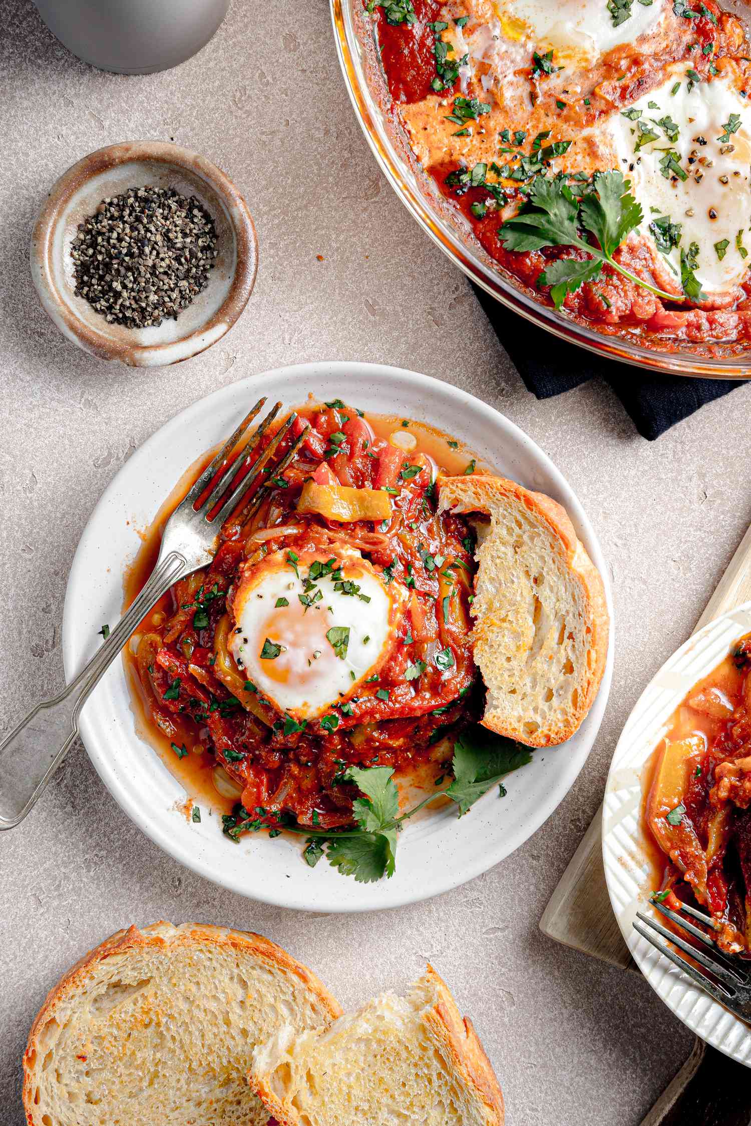 Serving of Microwave Shakshuka with a Piece of Toasted Bread on a Small Plate, and in the Surroundings, More Bread on the Counter, Another Serving of Shakshuka on Another Plate, a Small Saucer with Cracked Pepper, and More Shakshuka in a Pyrex Dish